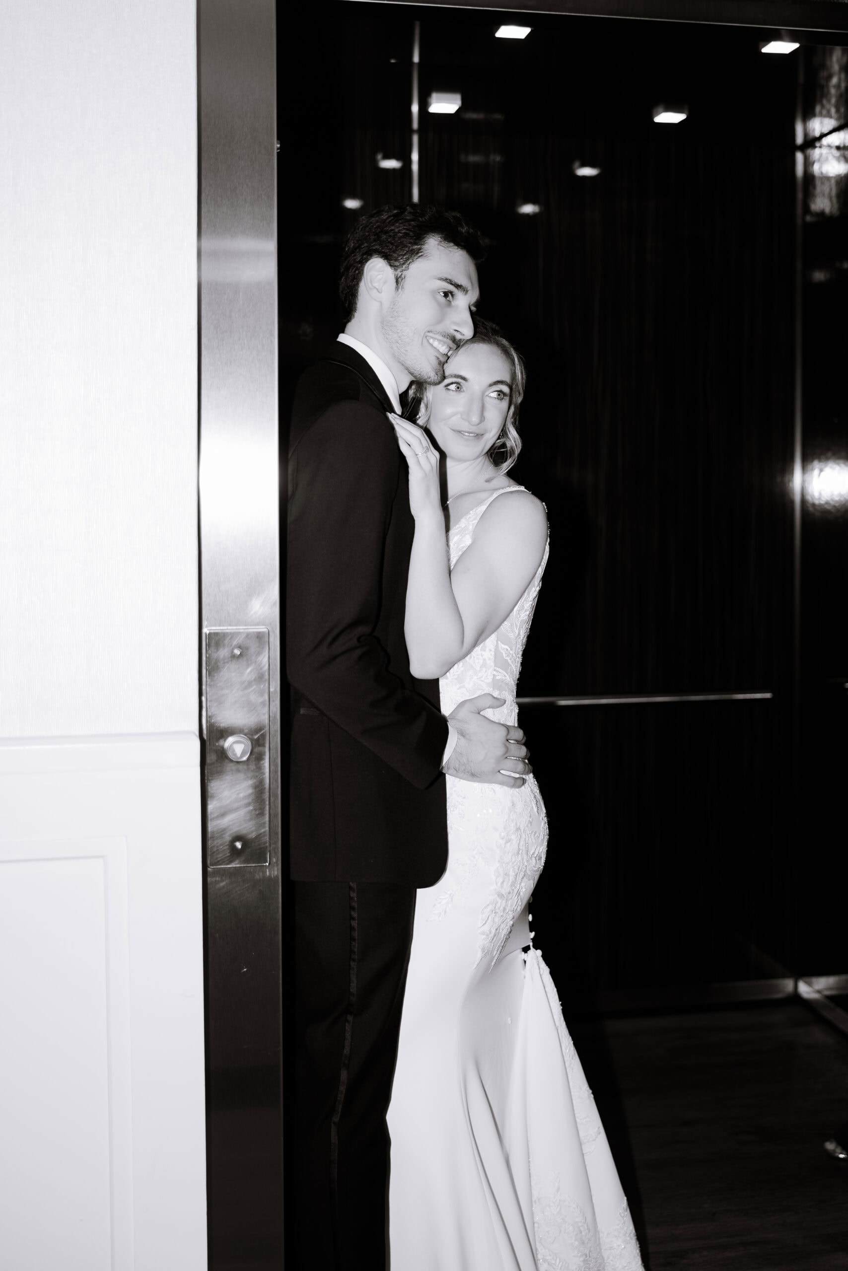Black and white photo of a groom in tuxedo embracing a bride in a lace wedding dress inside an elevator, highlighting textures and intimate connection.