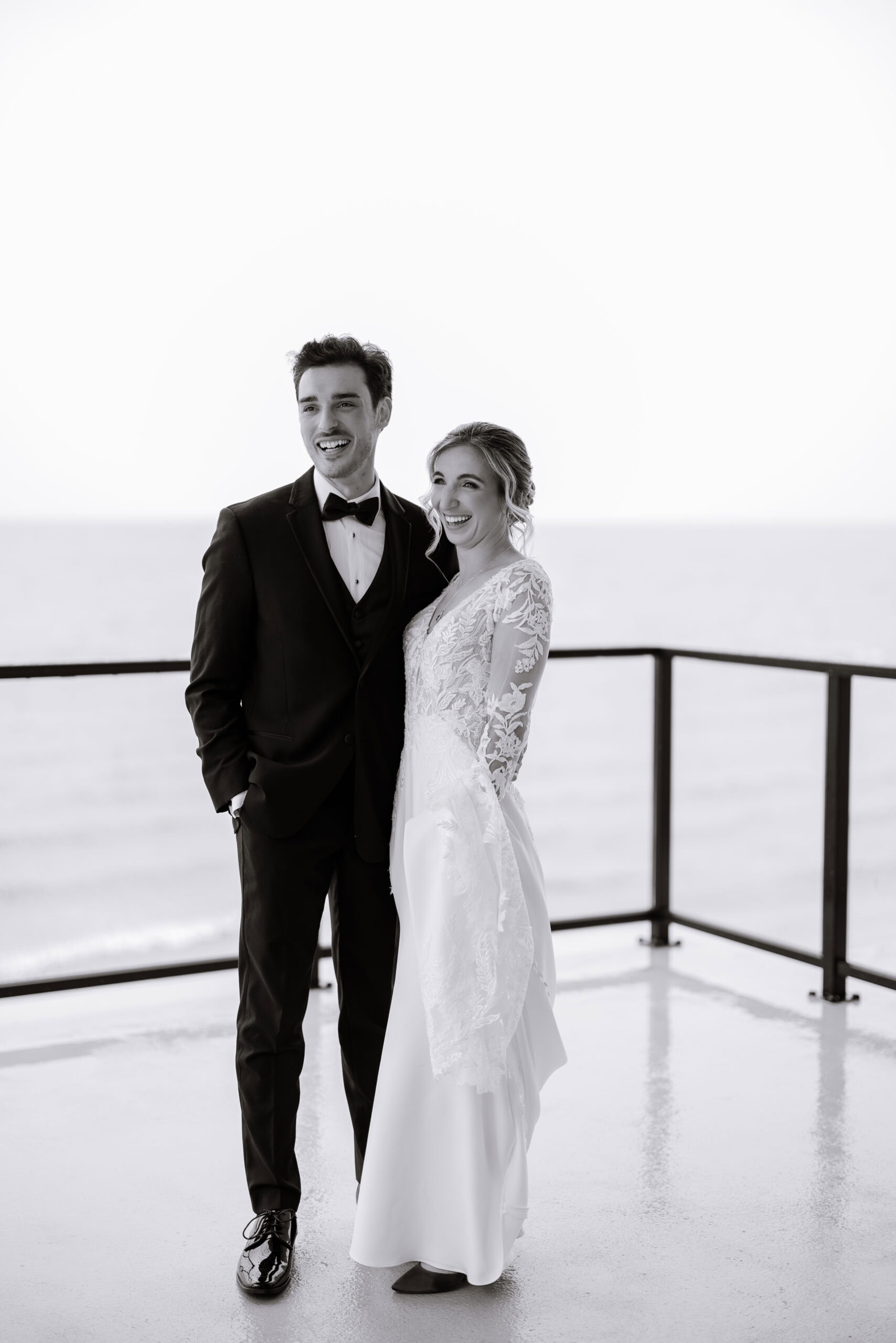 Black-and-white image of a couple in wedding attire smiling by a waterfront balcony at Tyde at Walnut Beach, Milford, CT.
