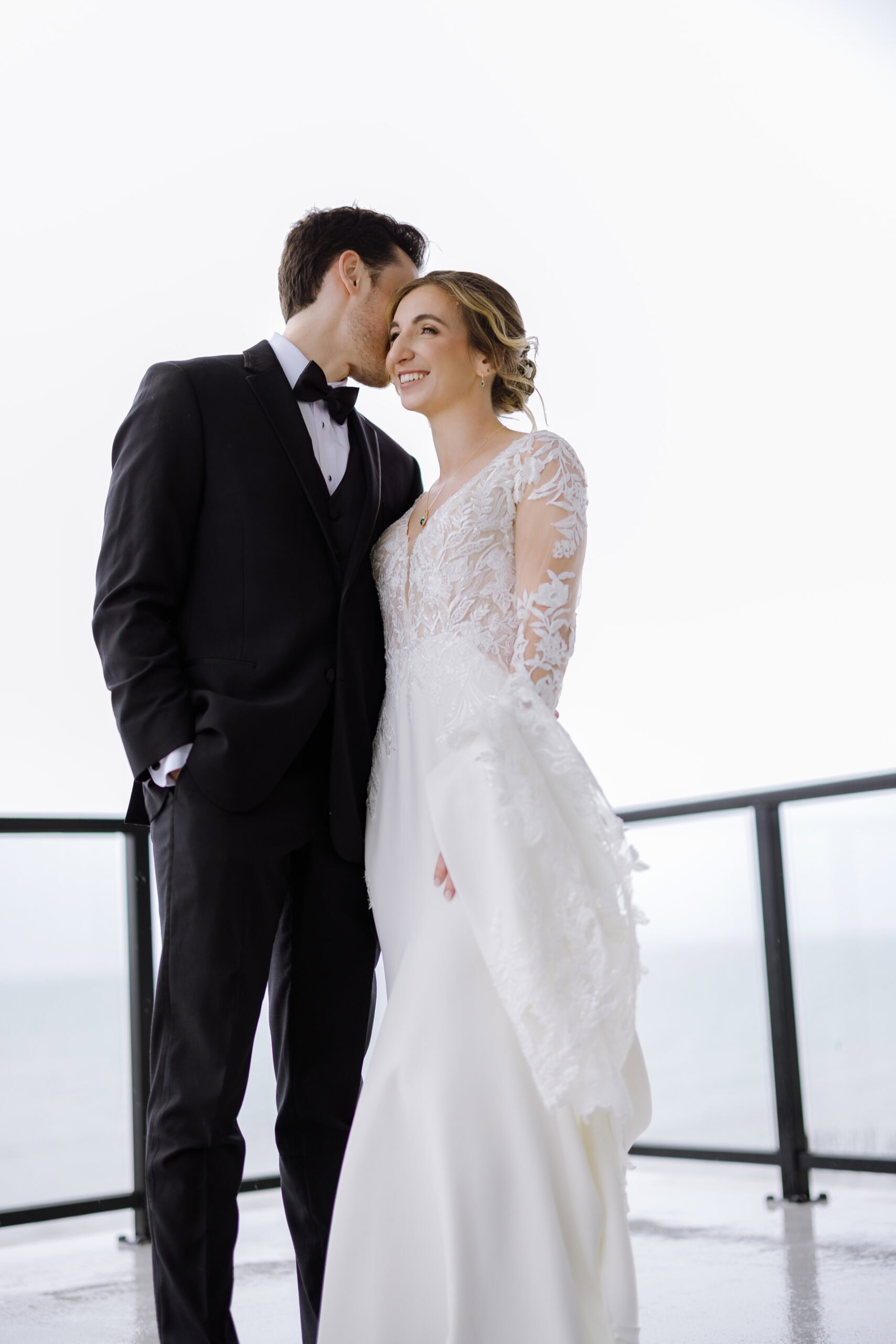 Groom in black tuxedo whispering to smiling bride in lace wedding gown on balcony overlooking water at Tyde at Walnut Beach, Milford, CT.