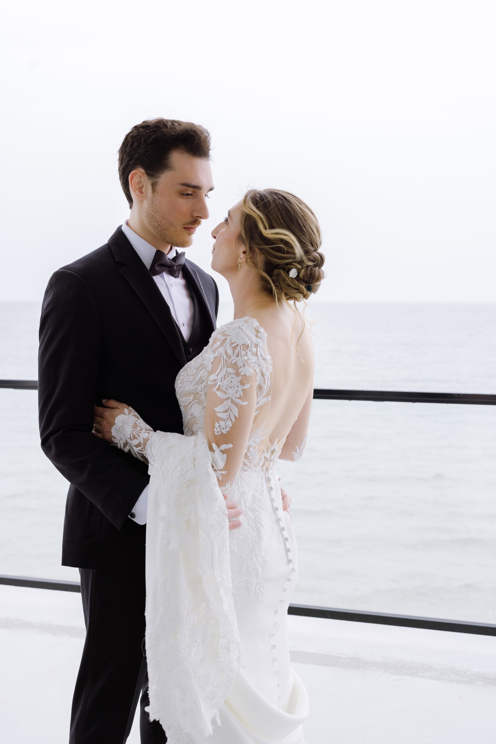 Bride and groom in elegant wedding attire embracing near a balcony overlooking the ocean at Tyde at Walnut Beach, Milford, CT.