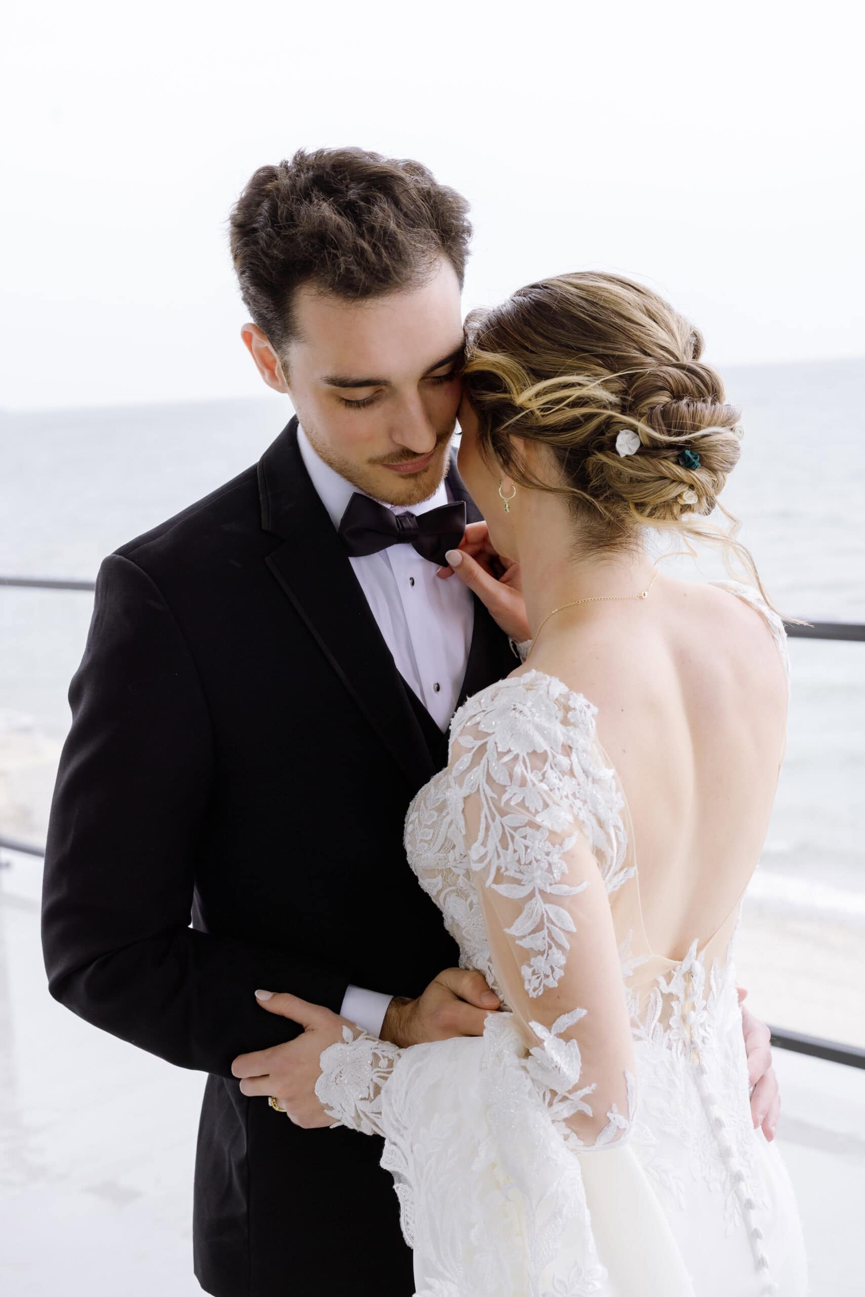 Bride and groom in an intimate embrace by the ocean, bride wearing lace gown with floral hairpins, groom in black tuxedo at Tyde at Walnut Beach