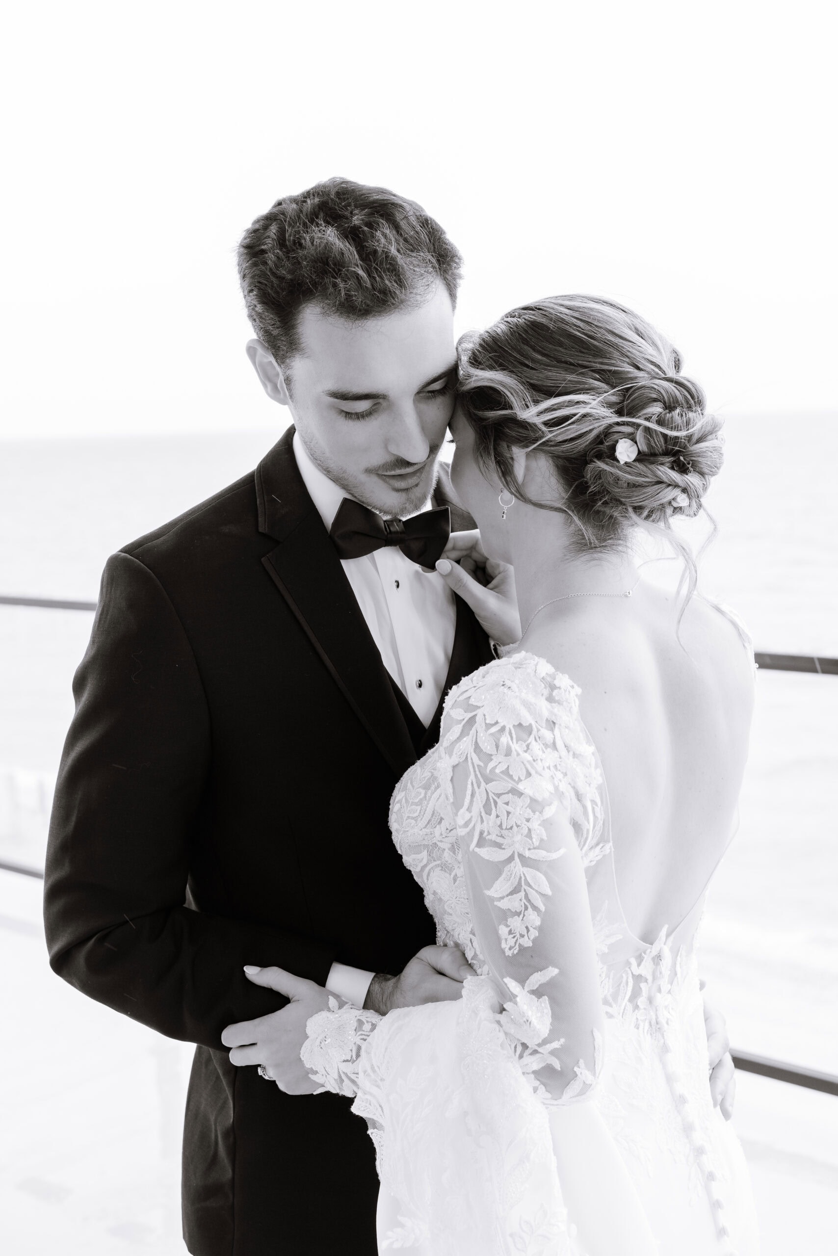 Bride in embroidered lace gown with open back embraces groom in black tuxedo adjusting his bow tie, ocean backdrop at Tyde at Walnut Beach, Milford CT.