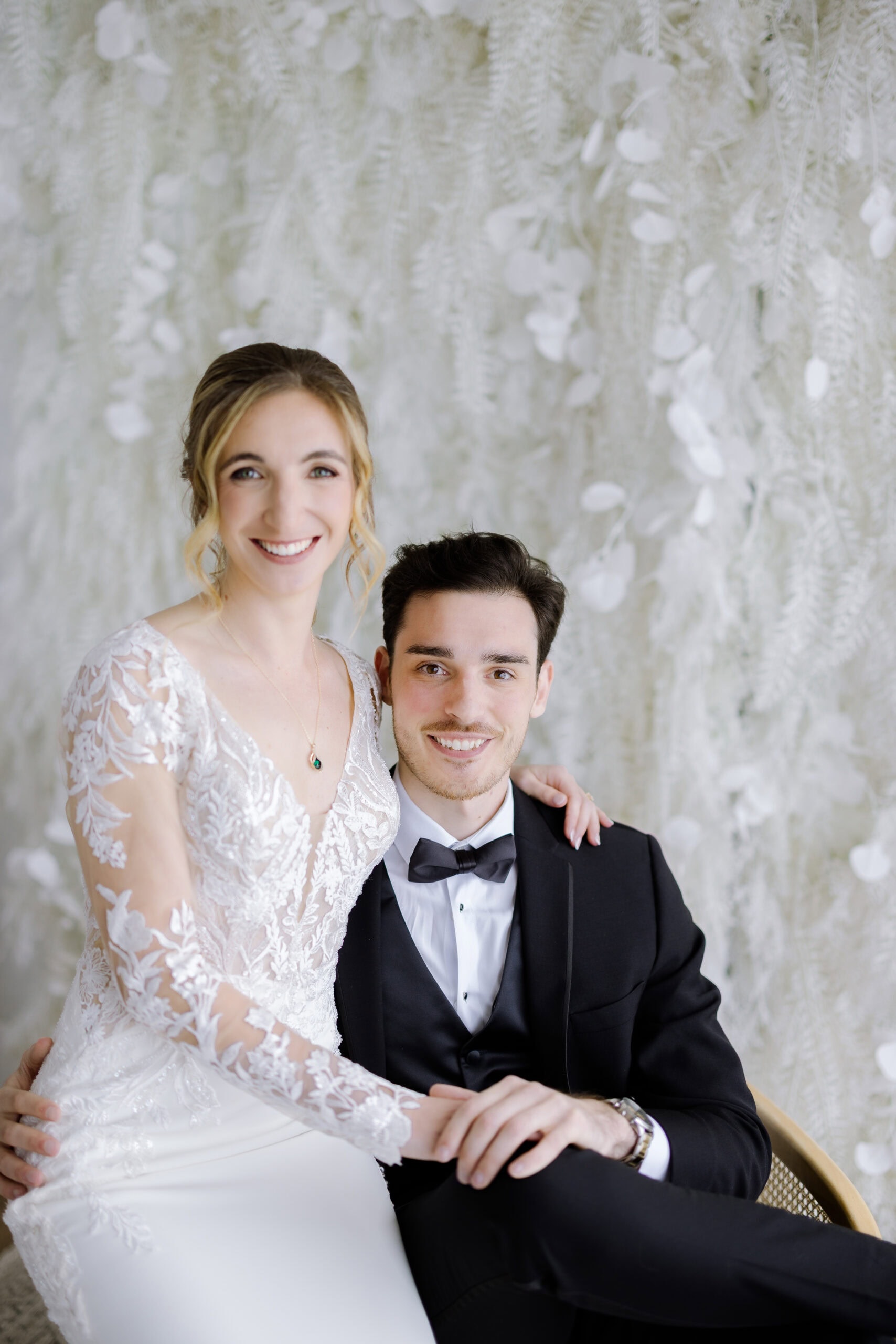 Bride in lace wedding dress with emerald pendant necklace and groom in black tuxedo seated together before a white textured botanical backdrop at Tyde at Walnut Beach, Milford, CT.