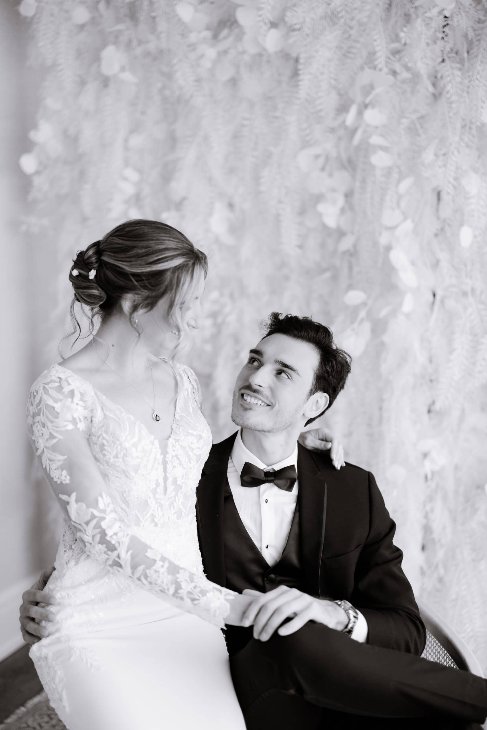 Black and white photo of bride in lace gown sitting on groom's lap, both gazing at each other with a soft botanical backdrop.