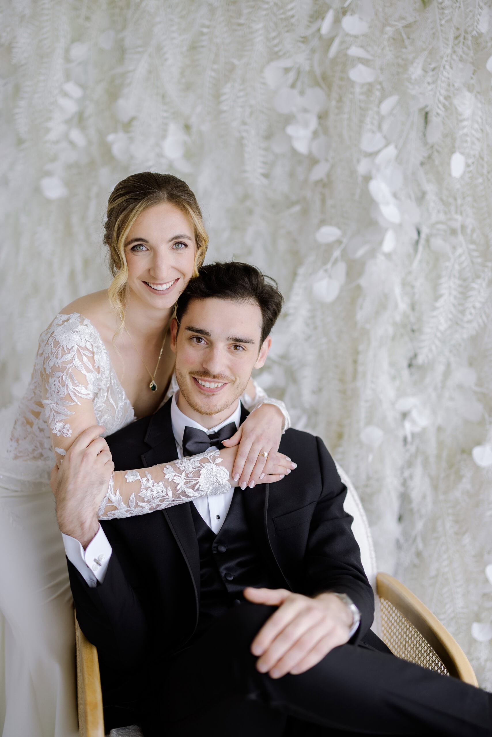 Bride in lace gown embraces groom in black tuxedo seated in chair, with delicate white fern and petal backdrop at Tyde at Walnut Beach, Milford, CT.