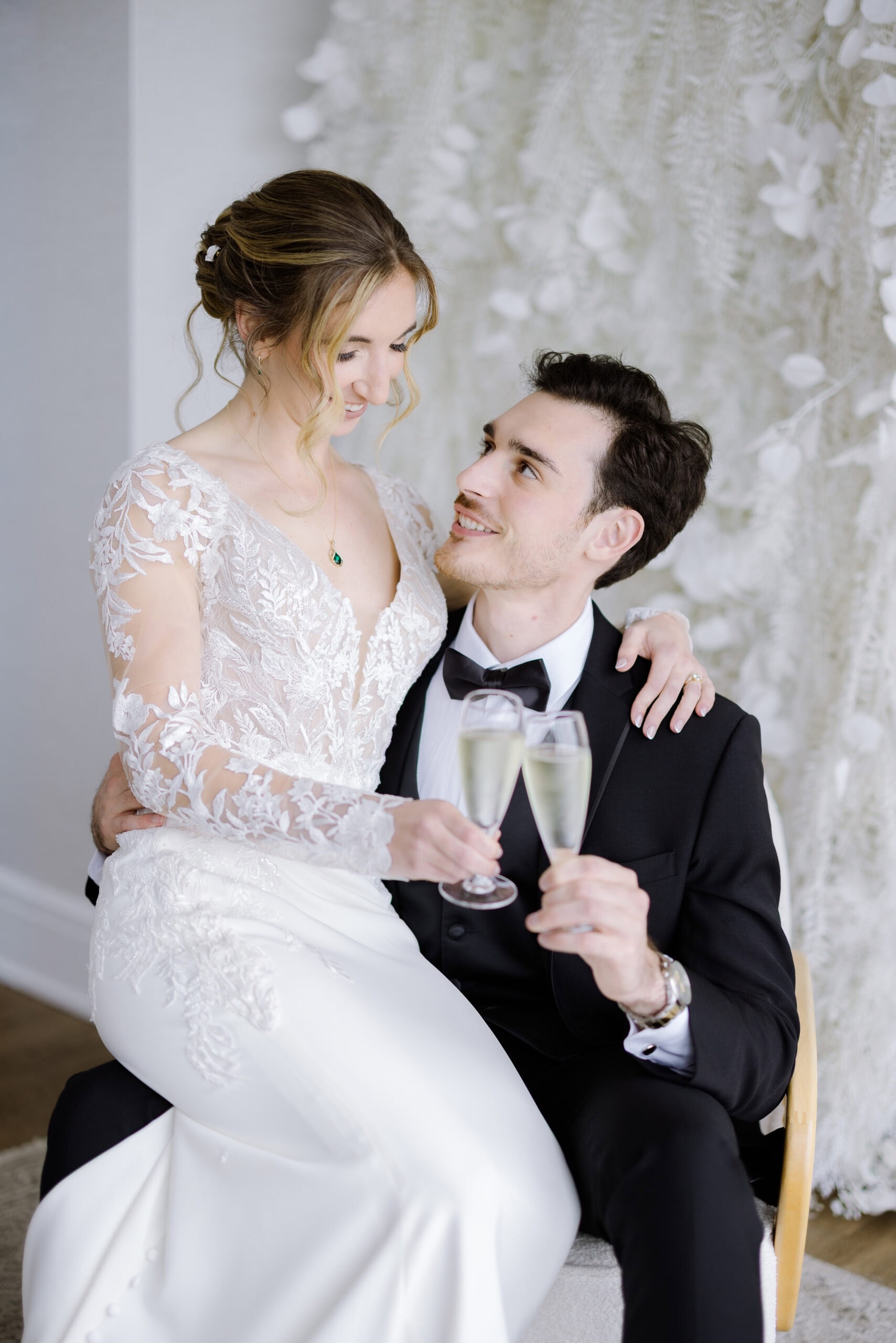 Bride in lace wedding dress sitting on groom's lap, both holding champagne glasses, with a textured white floral backdrop.