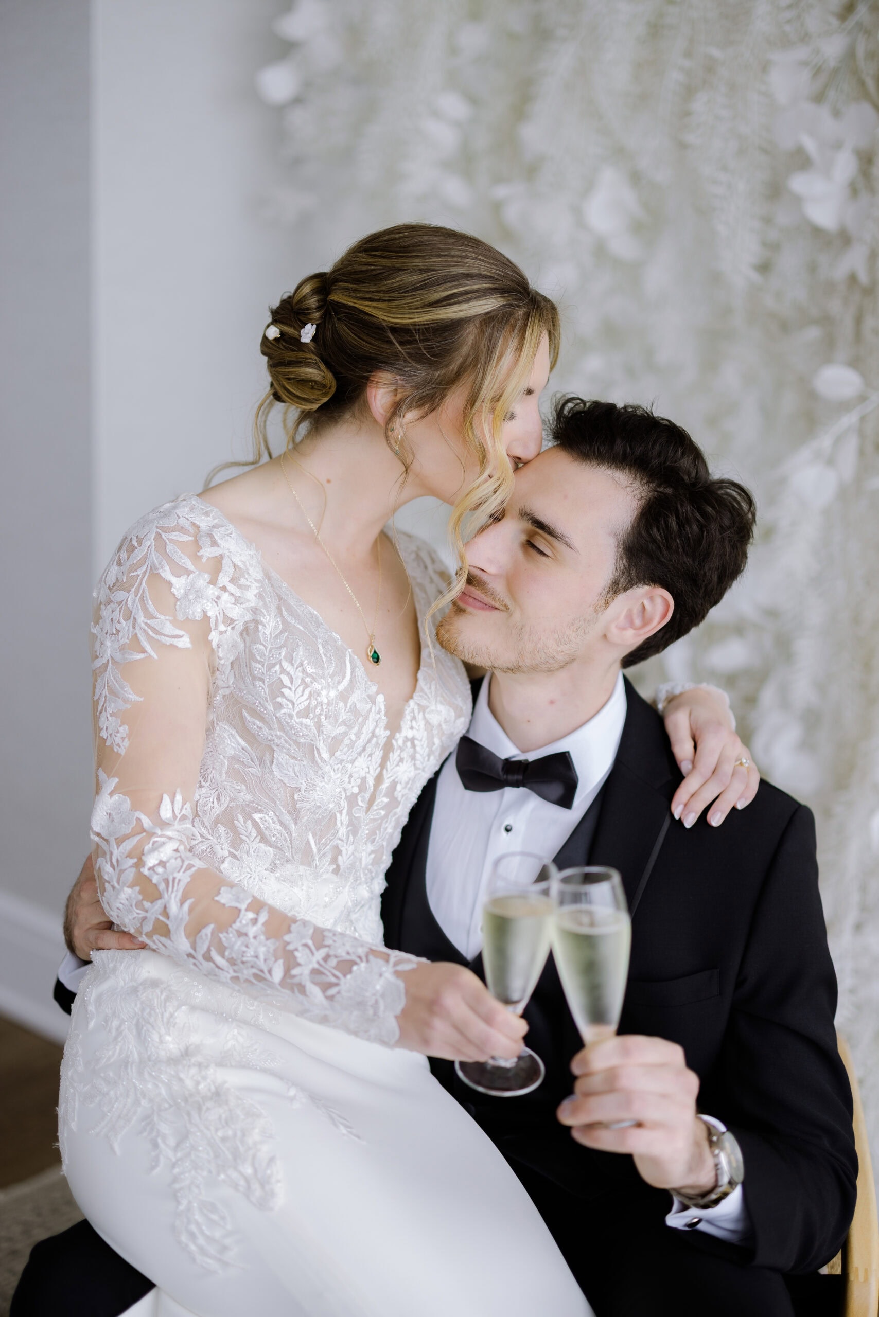 Bride in lace wedding dress kissing groom's forehead while holding champagne glasses, seated indoors with soft floral backdrop.