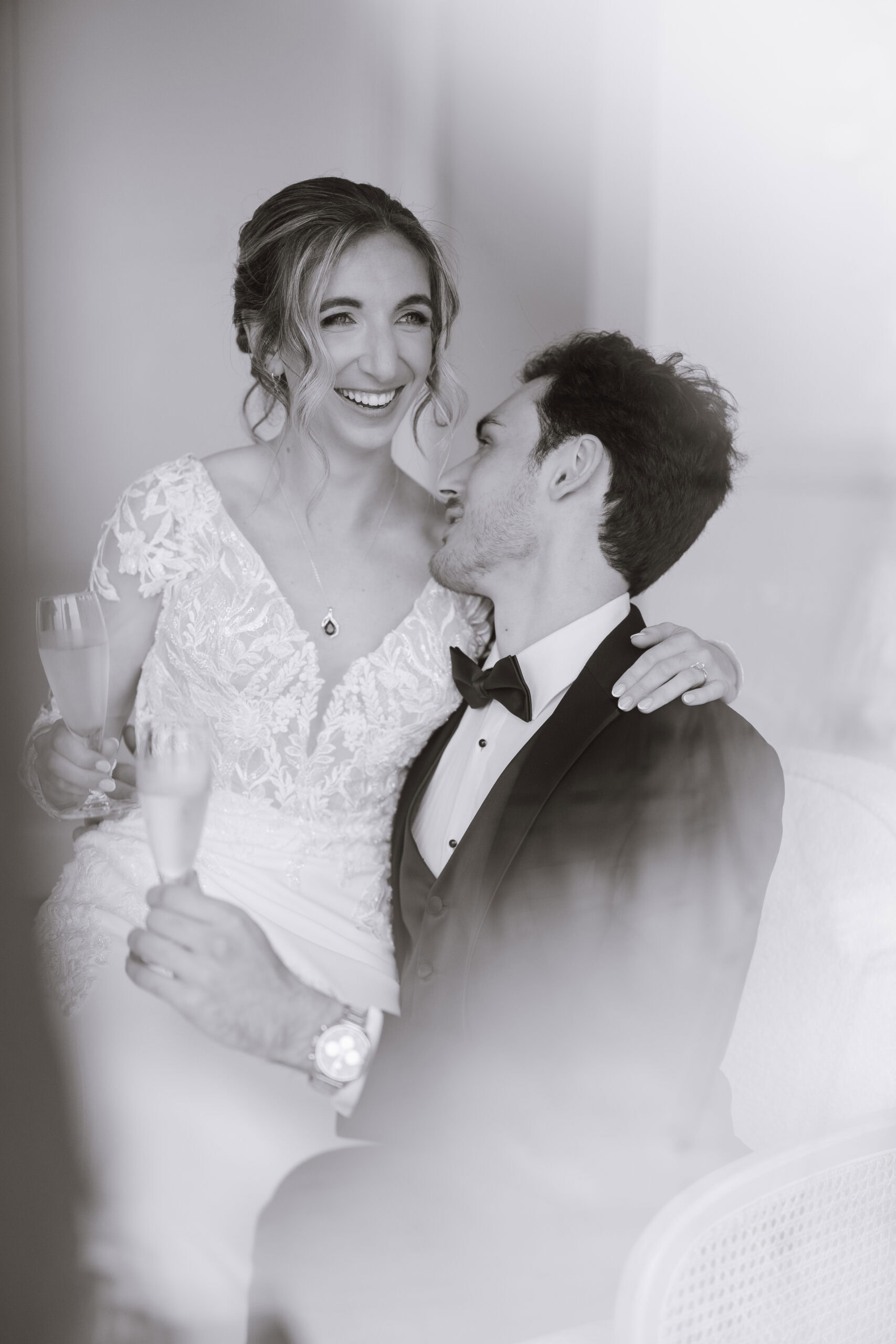 Black and white photo of a bride in a lace wedding dress smiling while sitting on the lap of a groom in a tuxedo, holding champagne flutes.