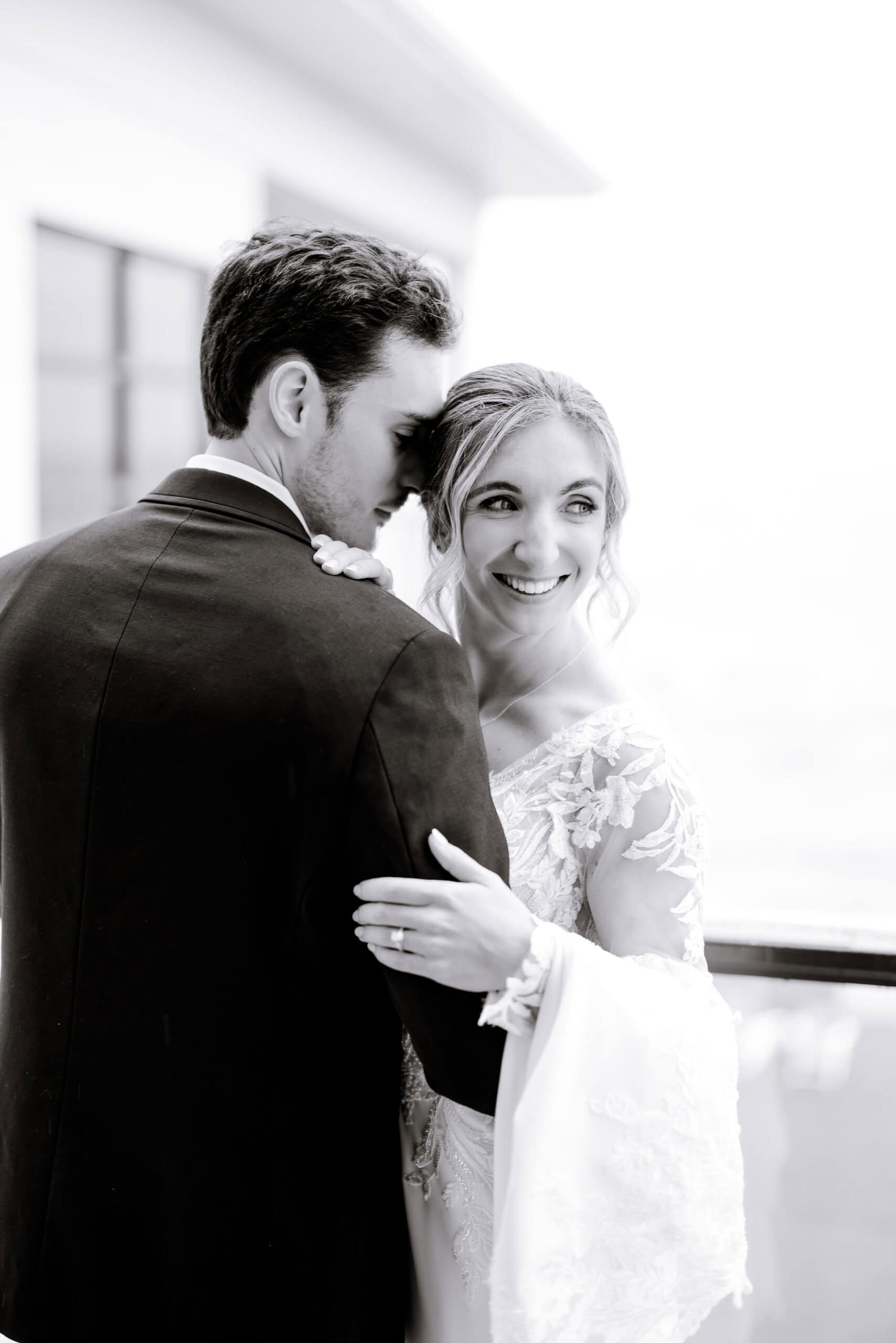 Black and white portrait of a bride in lace gown embracing groom in dark suit, soft focus background, Tyde at Walnut Beach venue.
