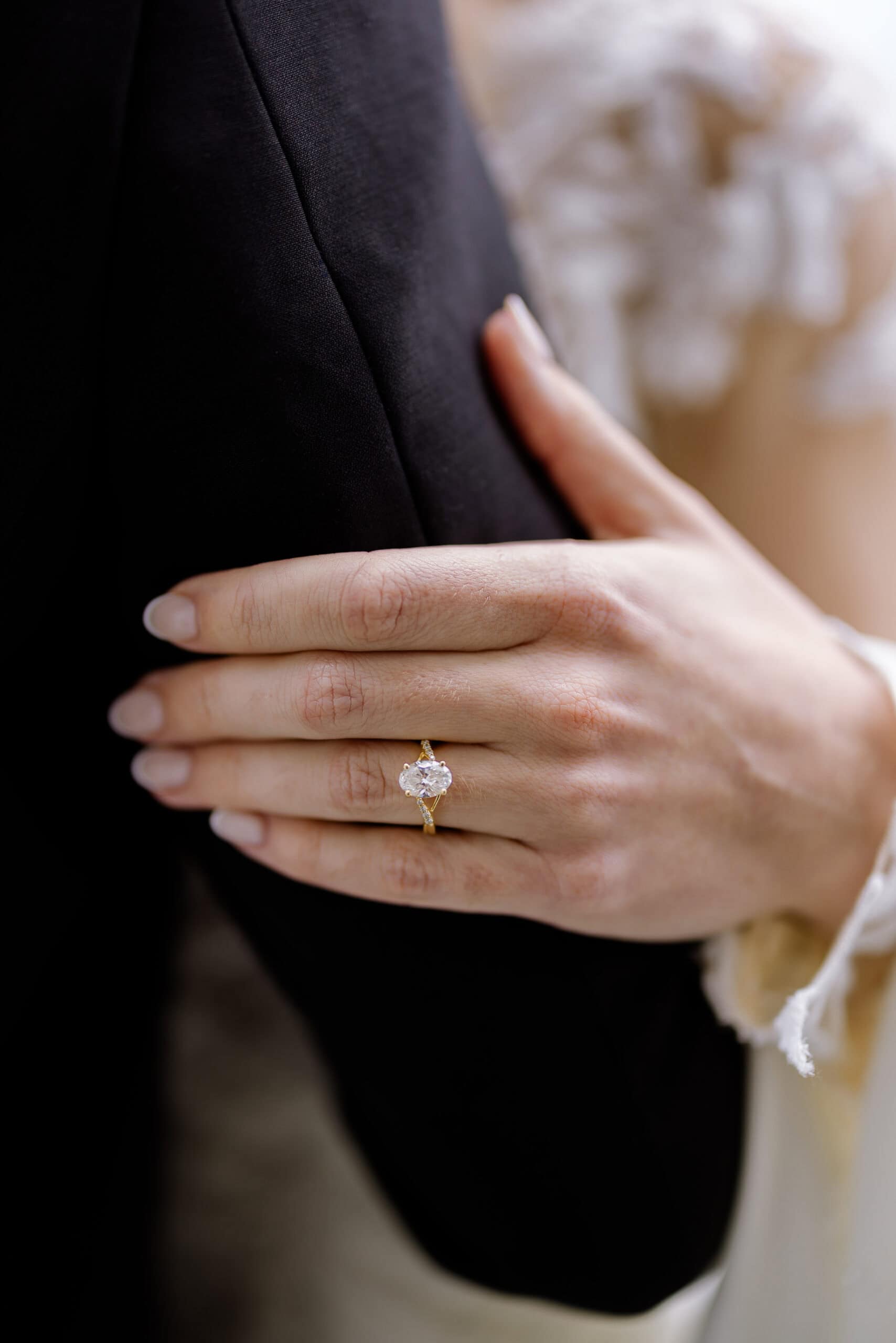 Close-up of a hand with a solitaire diamond engagement ring resting on a black suit jacket, showing fine fabric texture and soft lighting.