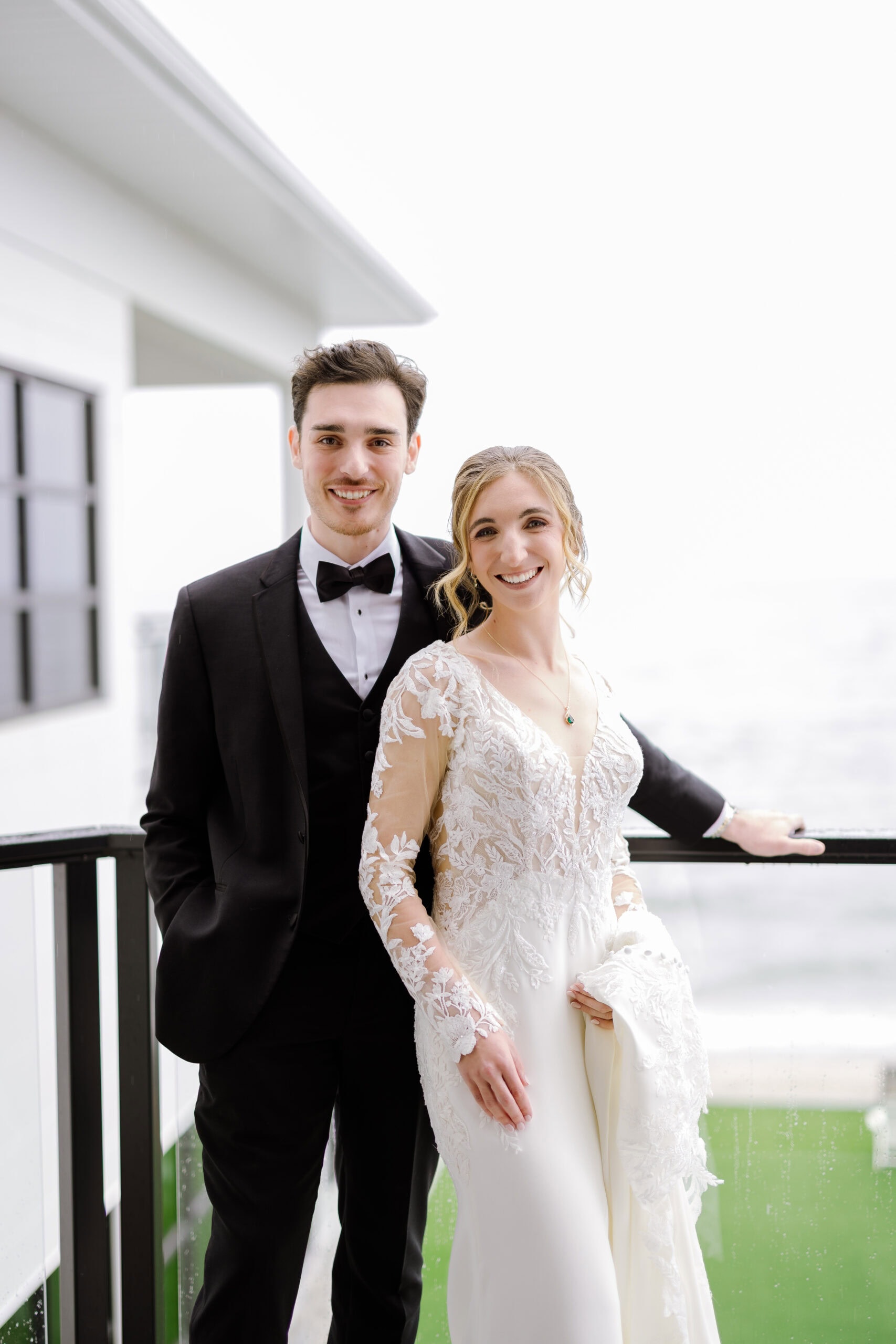 Bride in lace wedding dress and groom in black tuxedo standing on balcony with white architectural background and water view at Tyde at Walnut Beach, Milford, CT.