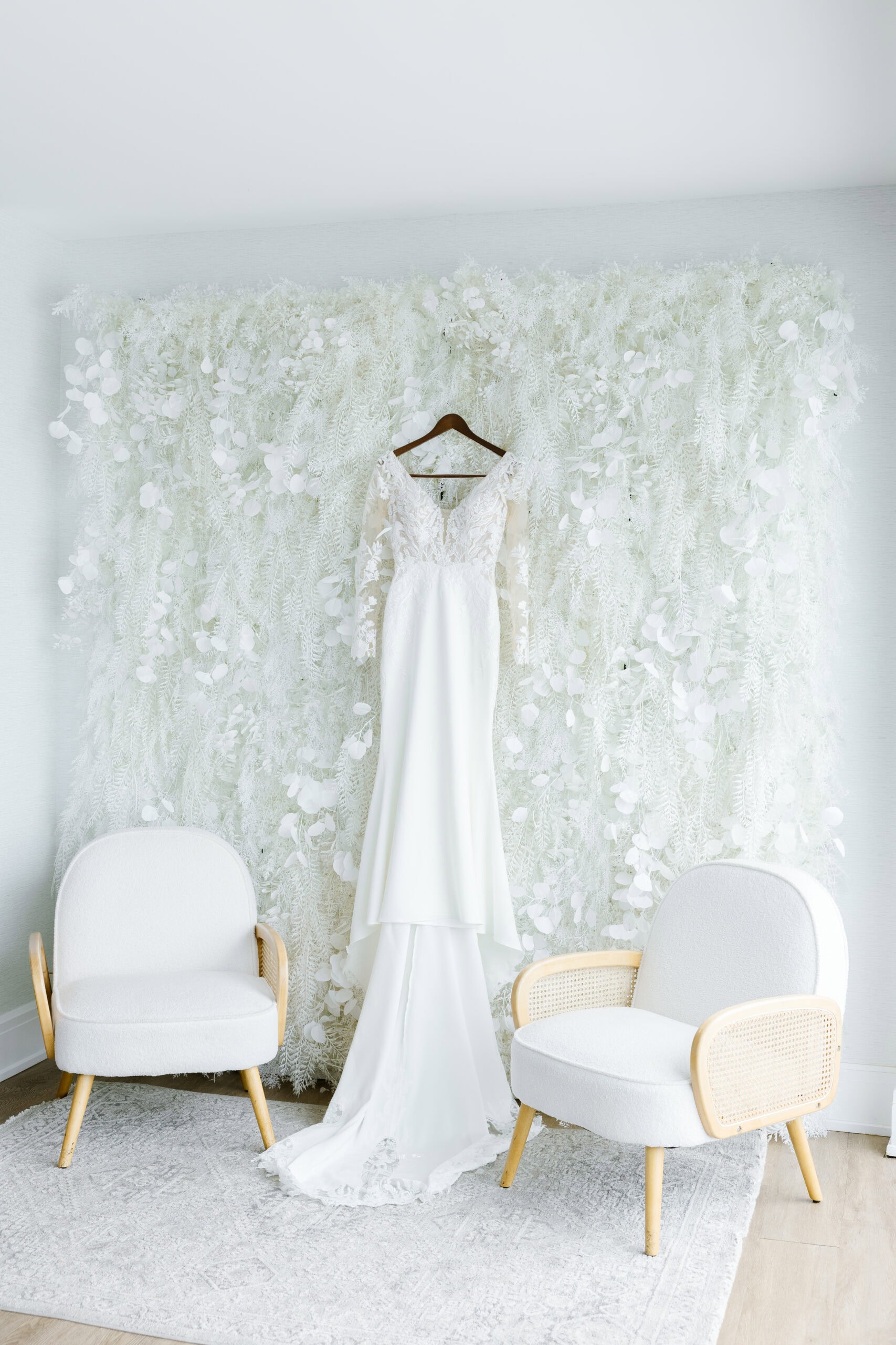 Long-sleeved lace wedding dress hanging on wooden hanger against white floral textured backdrop, flanked by two light-colored armchairs.
