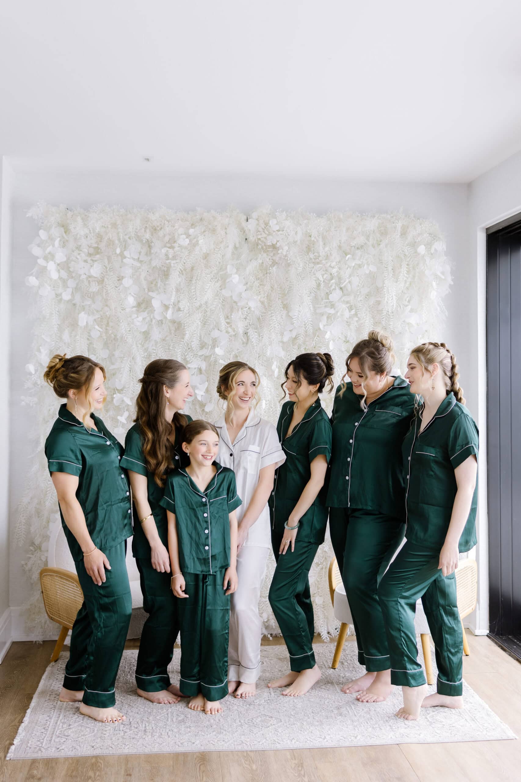 Bride in white silk pajamas with six bridesmaids in dark green silk pajamas standing barefoot on a rug, smiling and interacting indoors at Tyde at Walnut Beach, Milford, CT.
