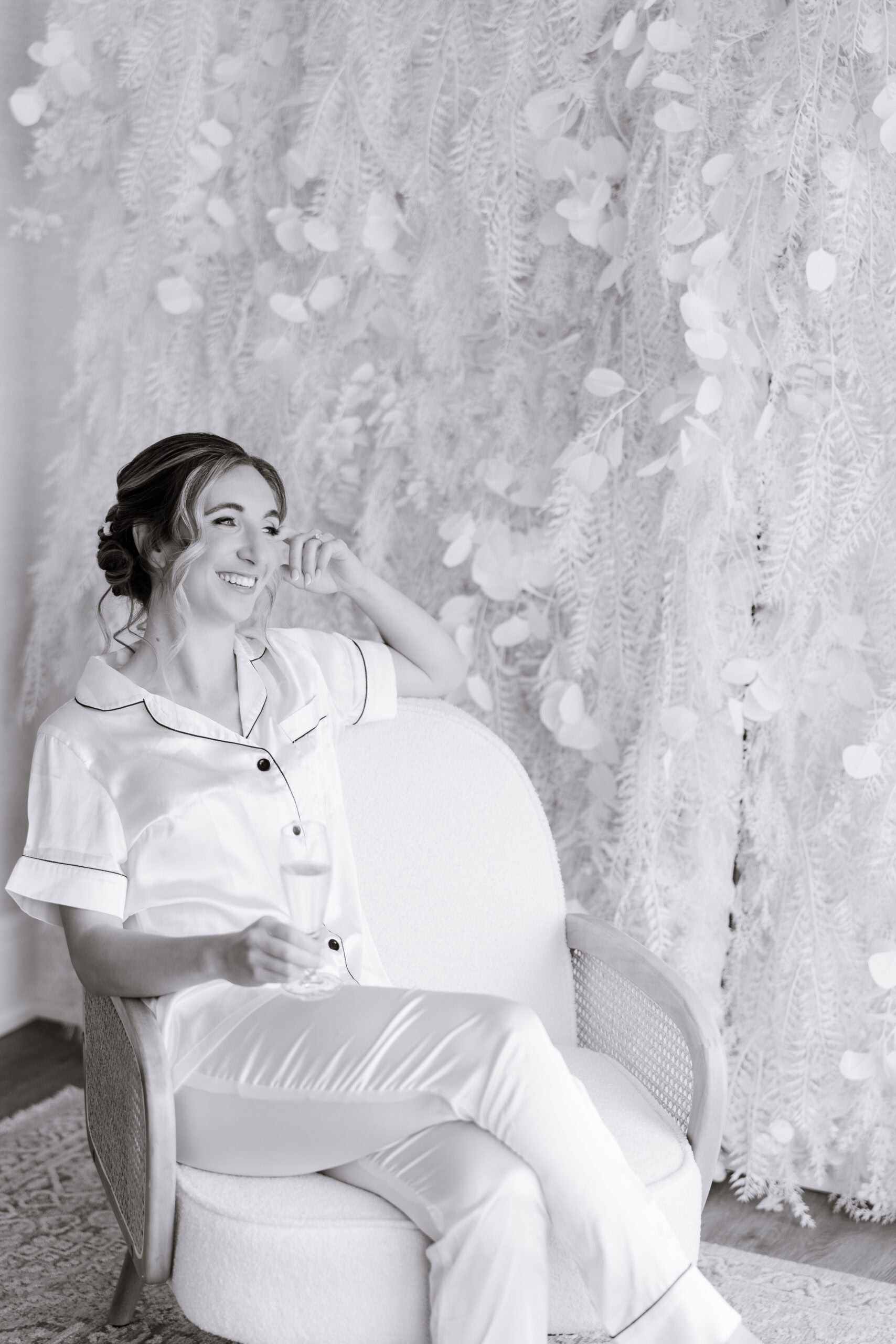 Bride seated in a white textured armchair wearing silk pajamas, holding a champagne flute, with a detailed white floral backdrop.