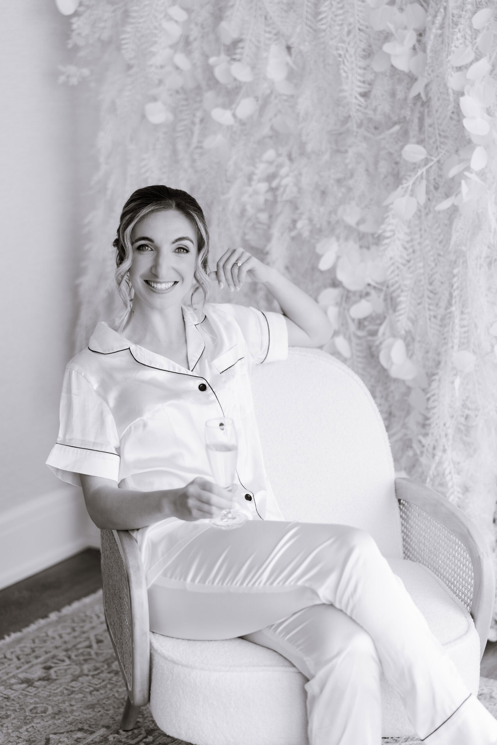 Bride seated in a textured armchair wearing silk pajamas, holding a champagne glass, with intricate floral backdrop at Tyde at Walnut Beach, Milford CT.