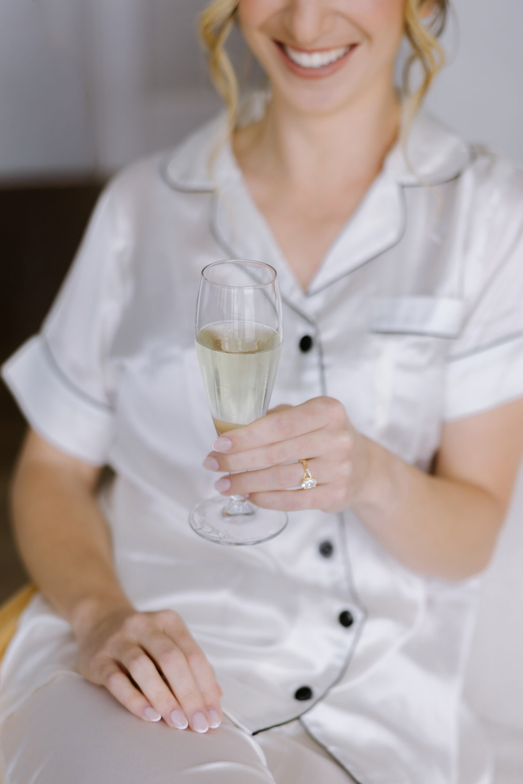 Close-up of a woman in silk pajamas holding a champagne flute, showcasing a pear-cut engagement ring and French manicure.