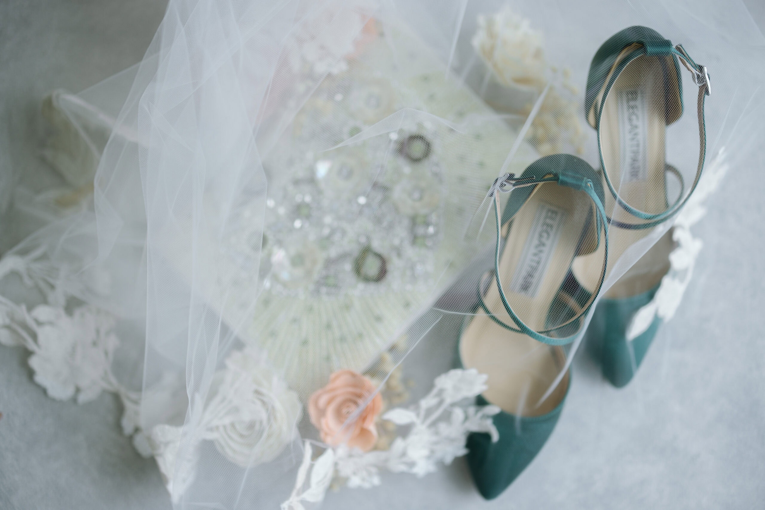 Top view of teal bridal heels partially covered by a white veil, with a detailed beaded bouquet featuring lace and soft flowers beneath.