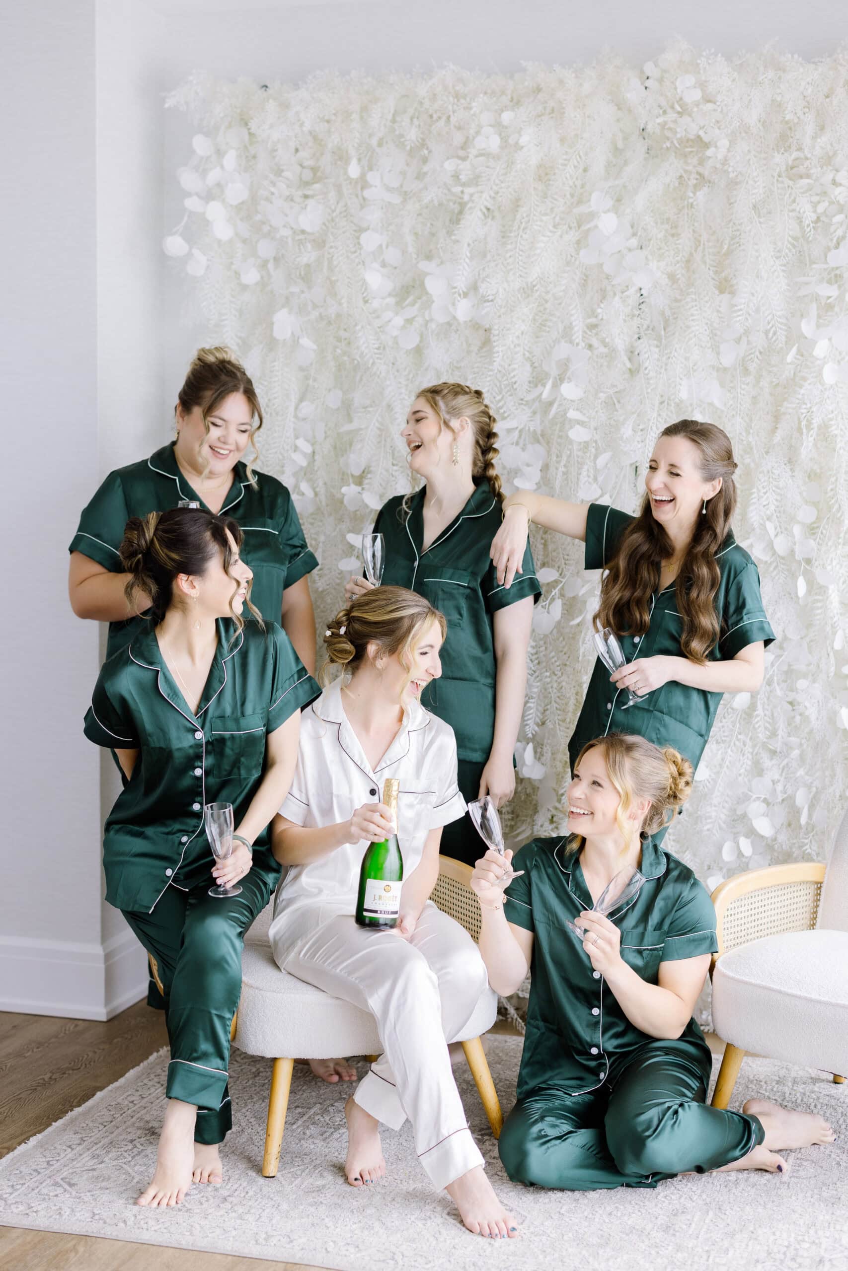 Bridal party in silk pajamas sharing champagne and laughter in front of a white floral wall at Tyde at Walnut Beach, Milford, CT.