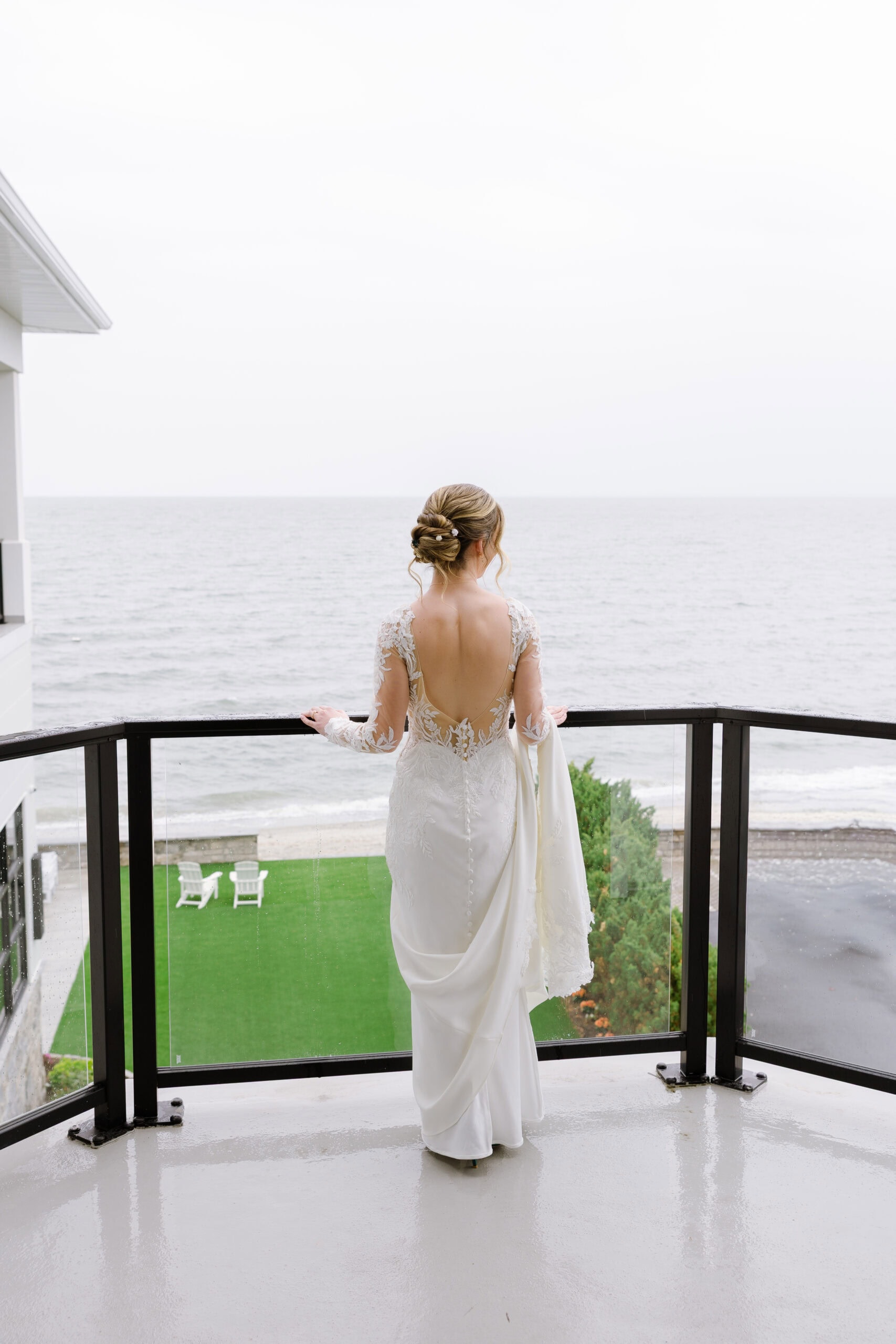 Bride in long-sleeved lace wedding dress with open back standing on balcony overlooking ocean and green lawn