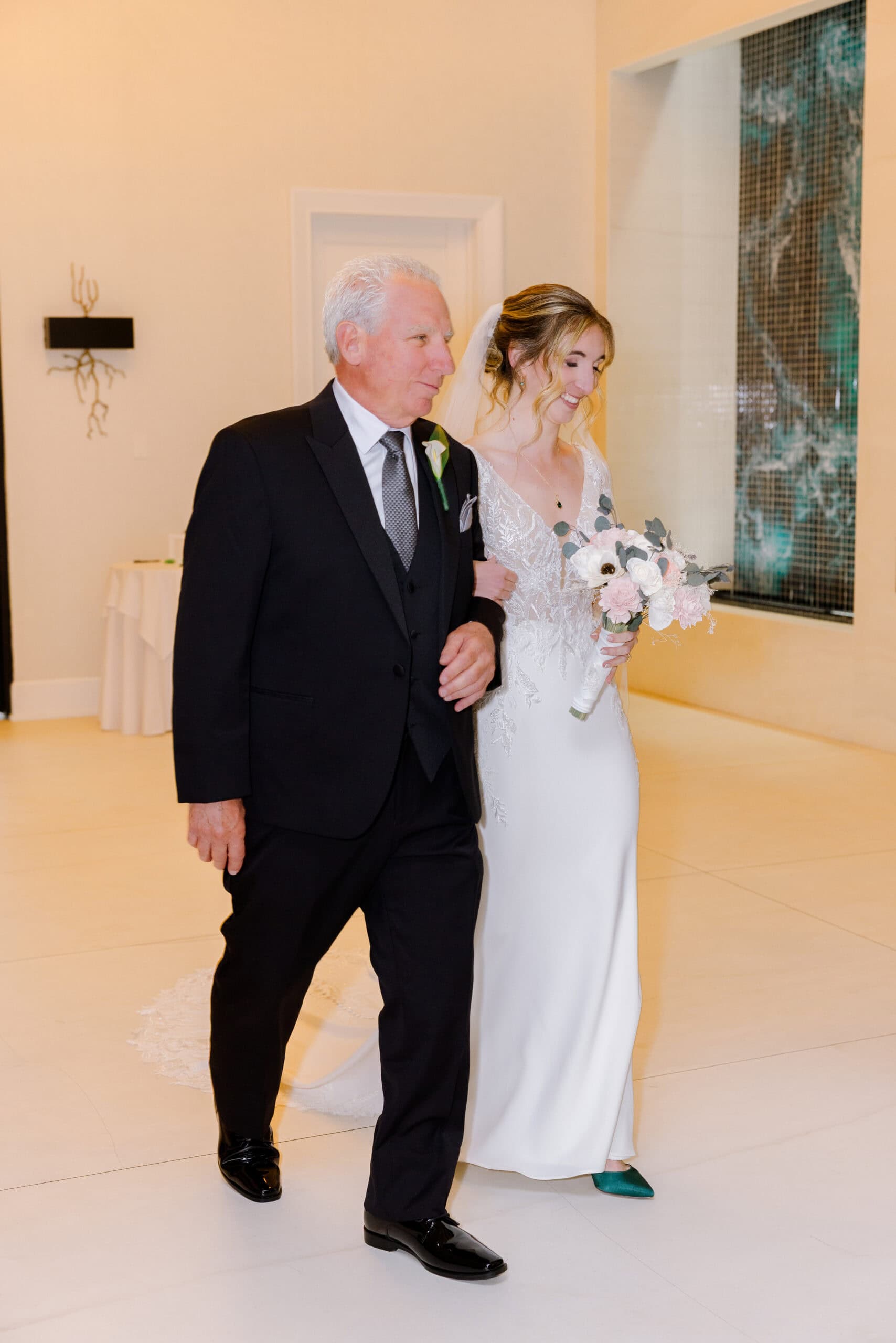 Bride in lace wedding dress holding bouquet walking arm-in-arm with father in black tuxedo inside a modern venue