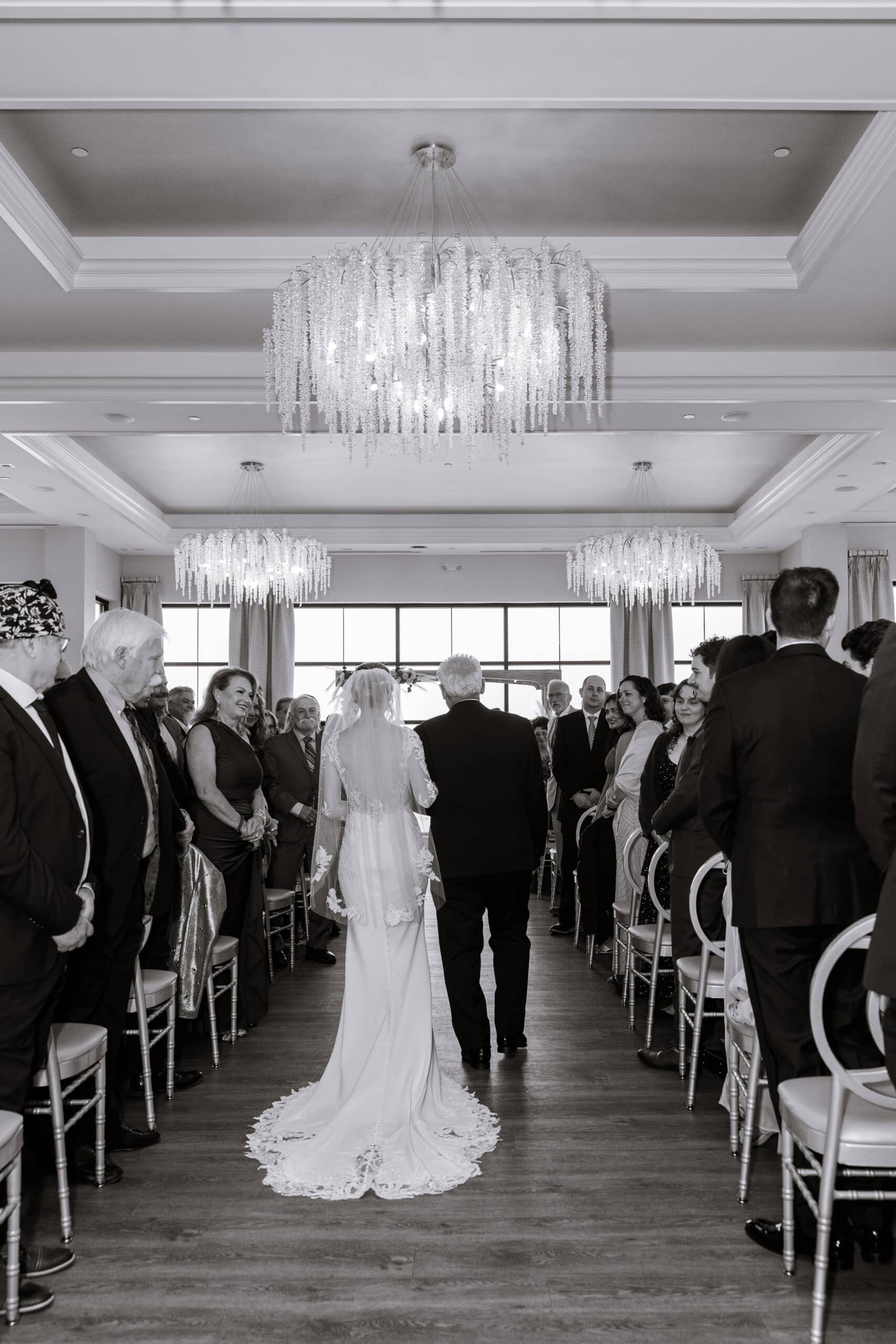 Bride walking down the aisle with escort under crystal chandeliers, guests seated on both sides in a bright venue at Tyde at Walnut Beach, Milford, CT.