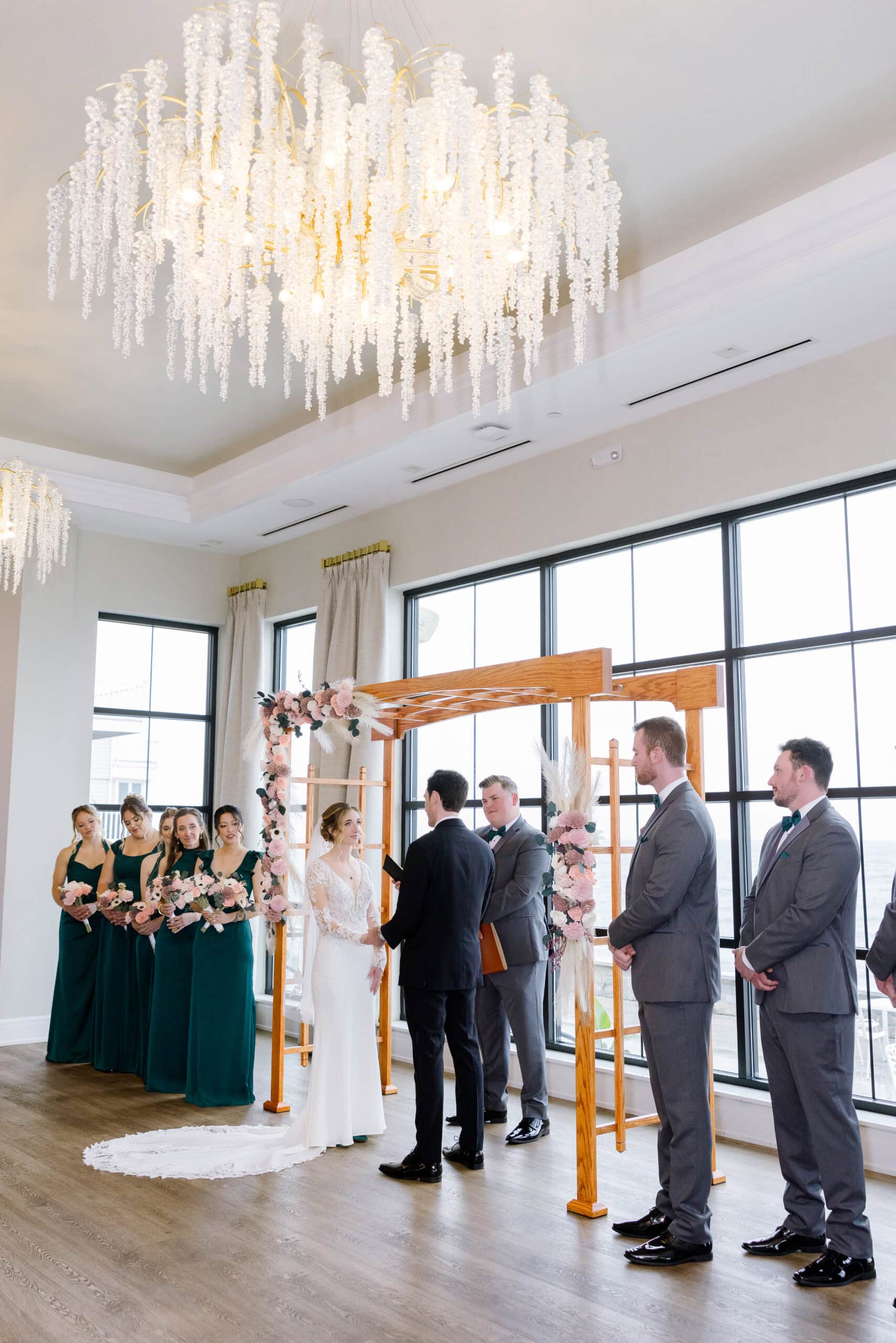 Bride and groom exchanging vows under a floral wooden arch with bridesmaids and groomsmen standing nearby beneath a large crystal chandelier at Tyde at Walnut Beach.