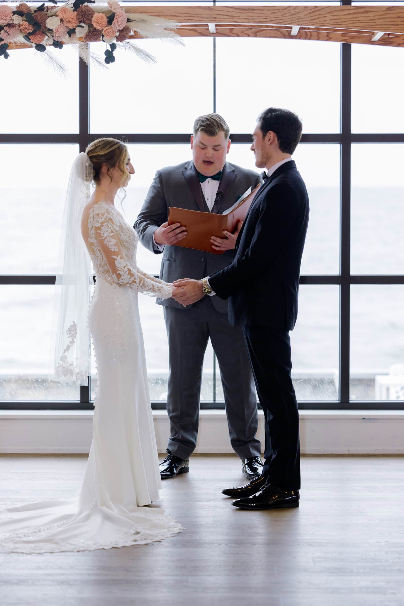 Bride in lace gown and groom in tuxedo holding hands during wedding ceremony with officiant reading vows in front of large window overlooking water at Tyde at Walnut Beach.