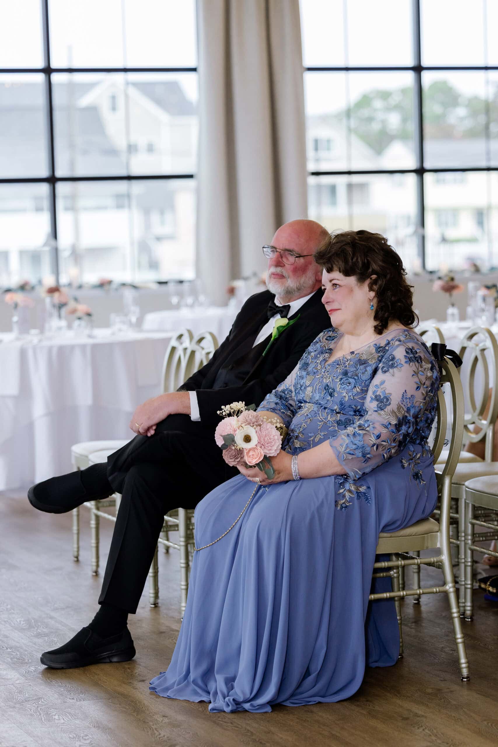 Seated older couple in formal attire at a wedding reception with large windows and floral decor at Tyde at Walnut Beach, Milford, CT.