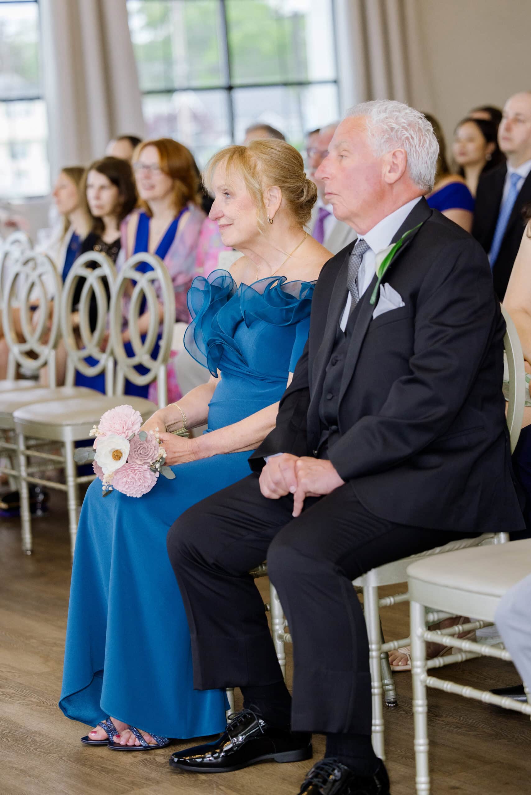 Older couple seated at wedding ceremony, woman in blue dress holding bouquet, man in black suit, guests blurred in background at Tyde at Walnut Beach, Milford, CT.