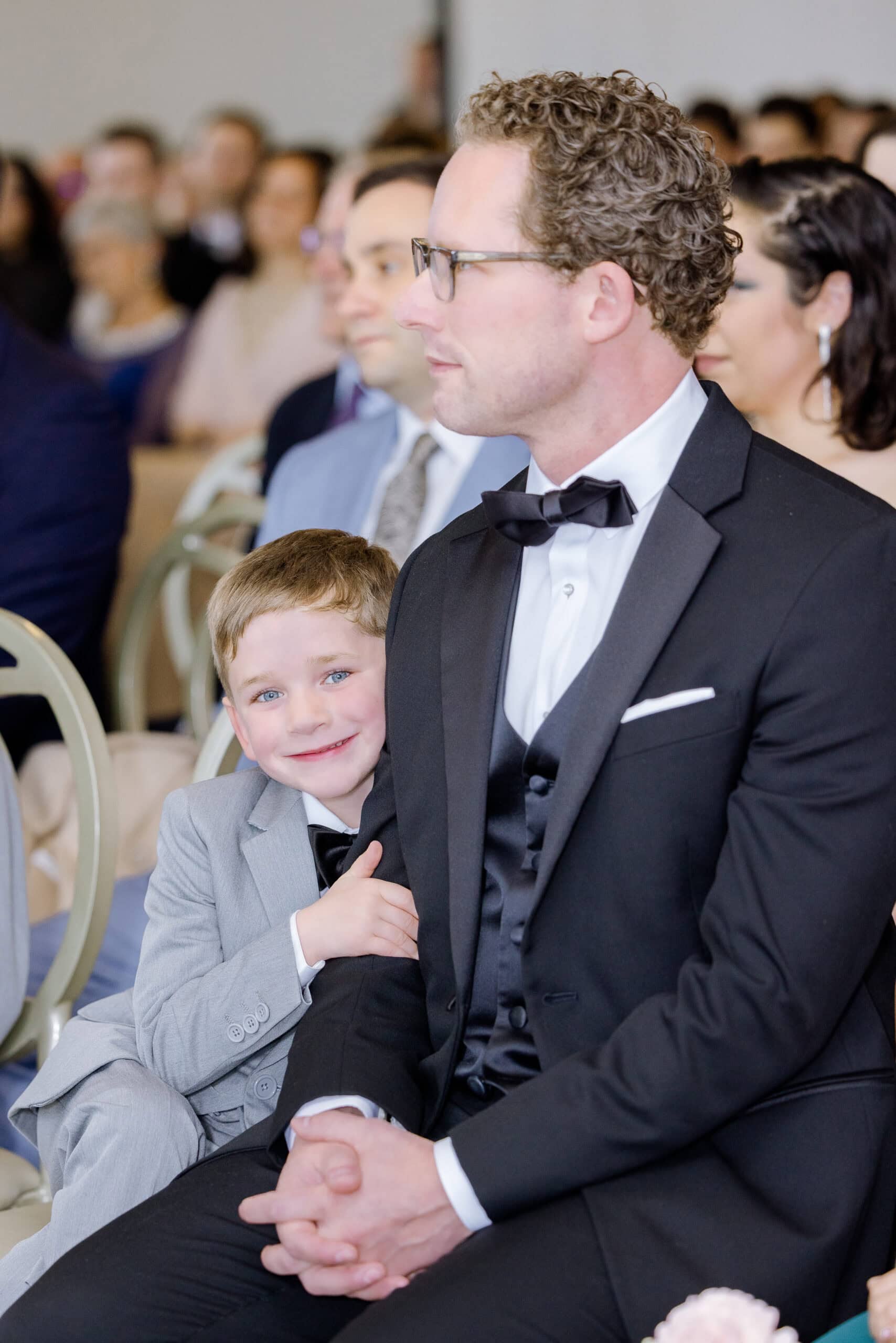 Young boy in gray suit smiling and hugging a man in black tuxedo seated at a wedding ceremony with blurred guests in the background.