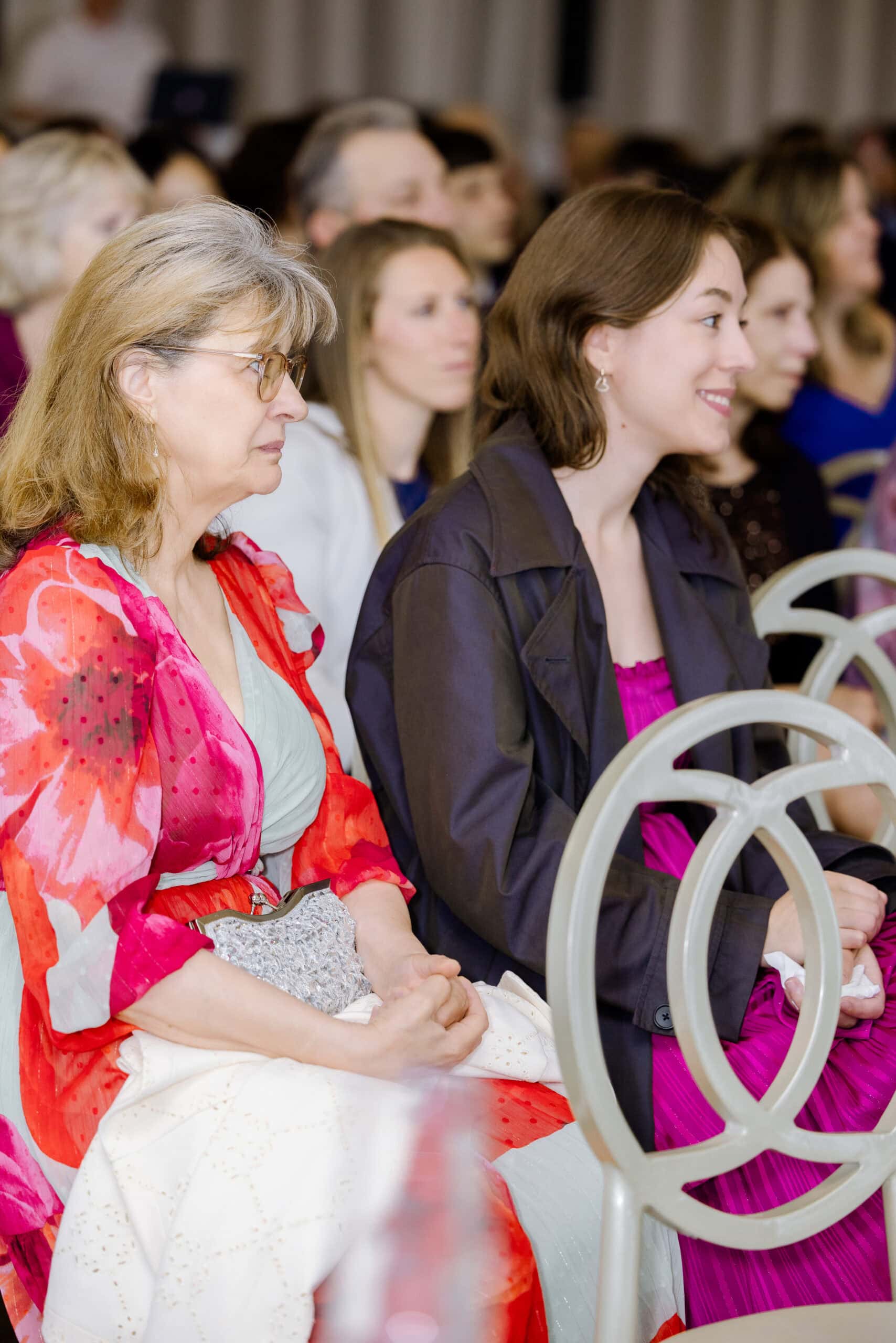Side view of two women seated in a crowd at a wedding, one in a red patterned shawl and glasses, the other in a dark jacket and pink dress, attentively watching the event.