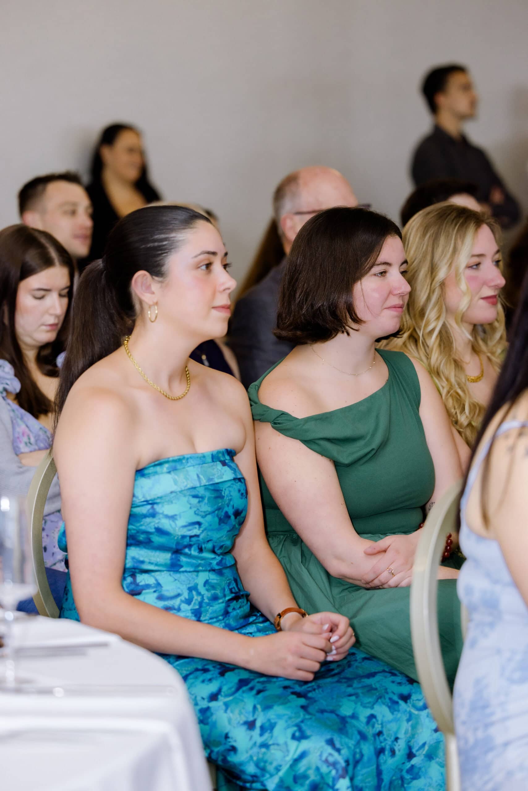Three women seated side by side at a wedding reception, dressed in elegant blue and green dresses, attentively watching the event.