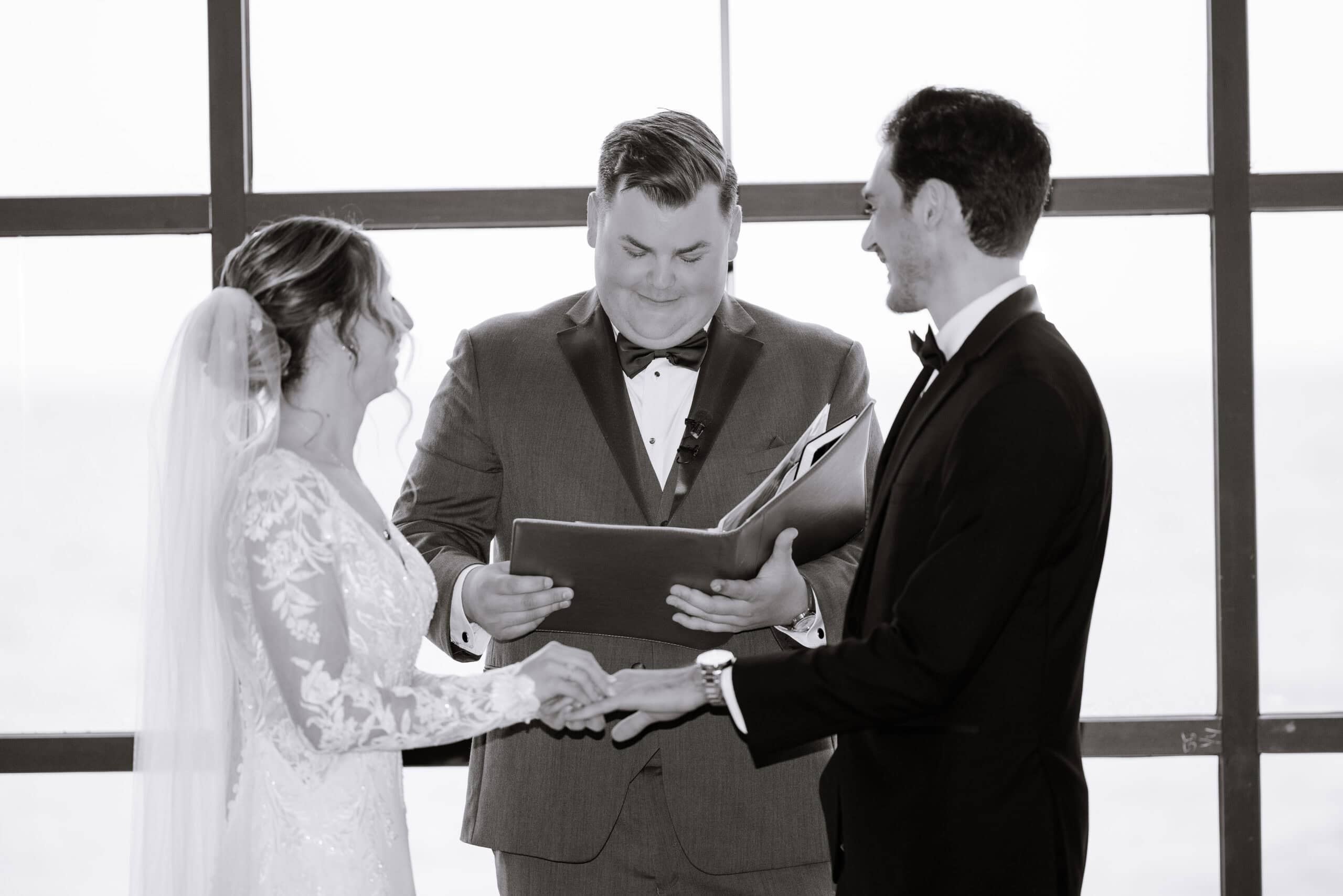 Bride and groom holding hands during wedding vows with officiant reading from book in front of large window.