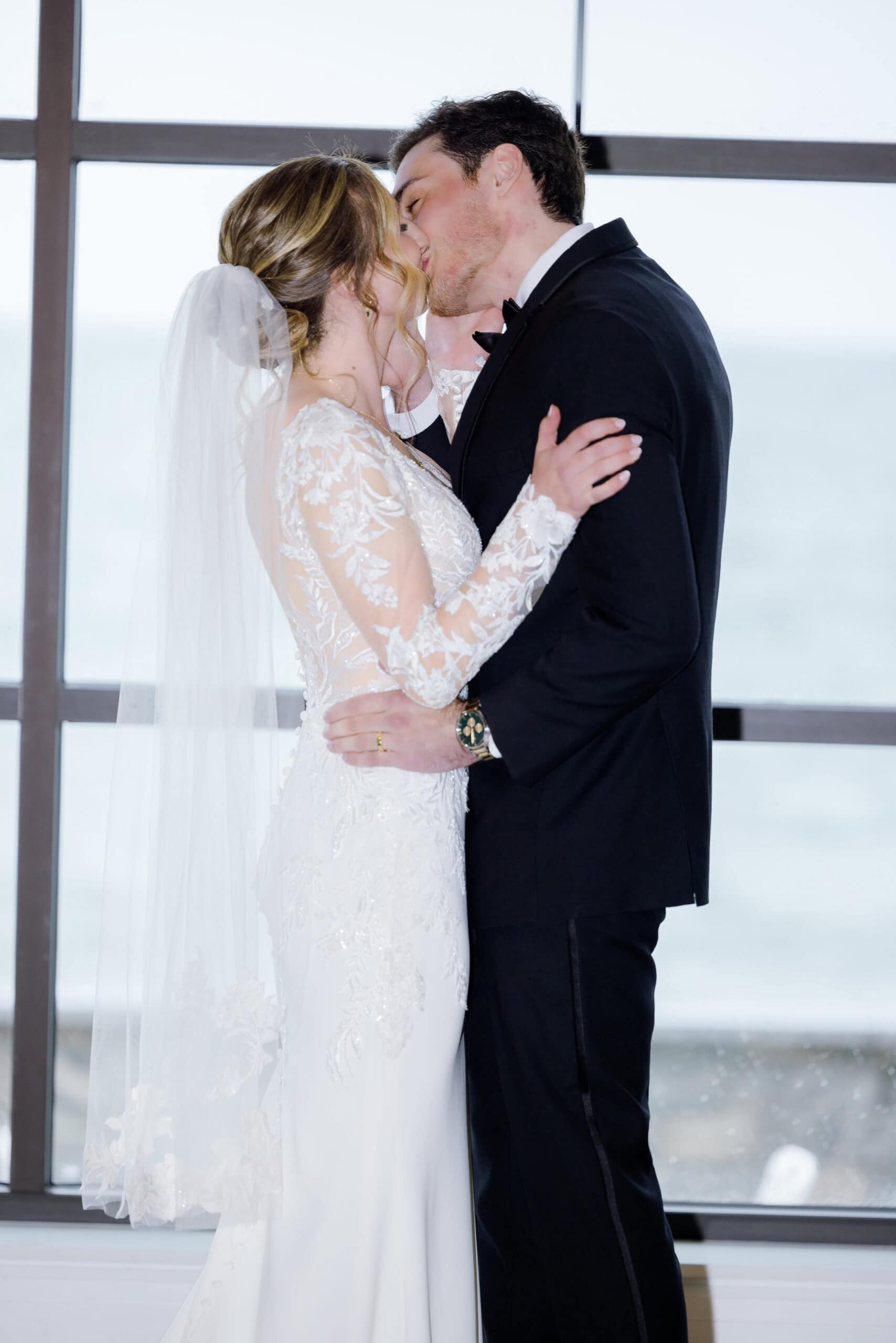 Bride and groom sharing a kiss in front of large windows with ocean view, bride wearing lace wedding dress and veil, groom in black tuxedo.