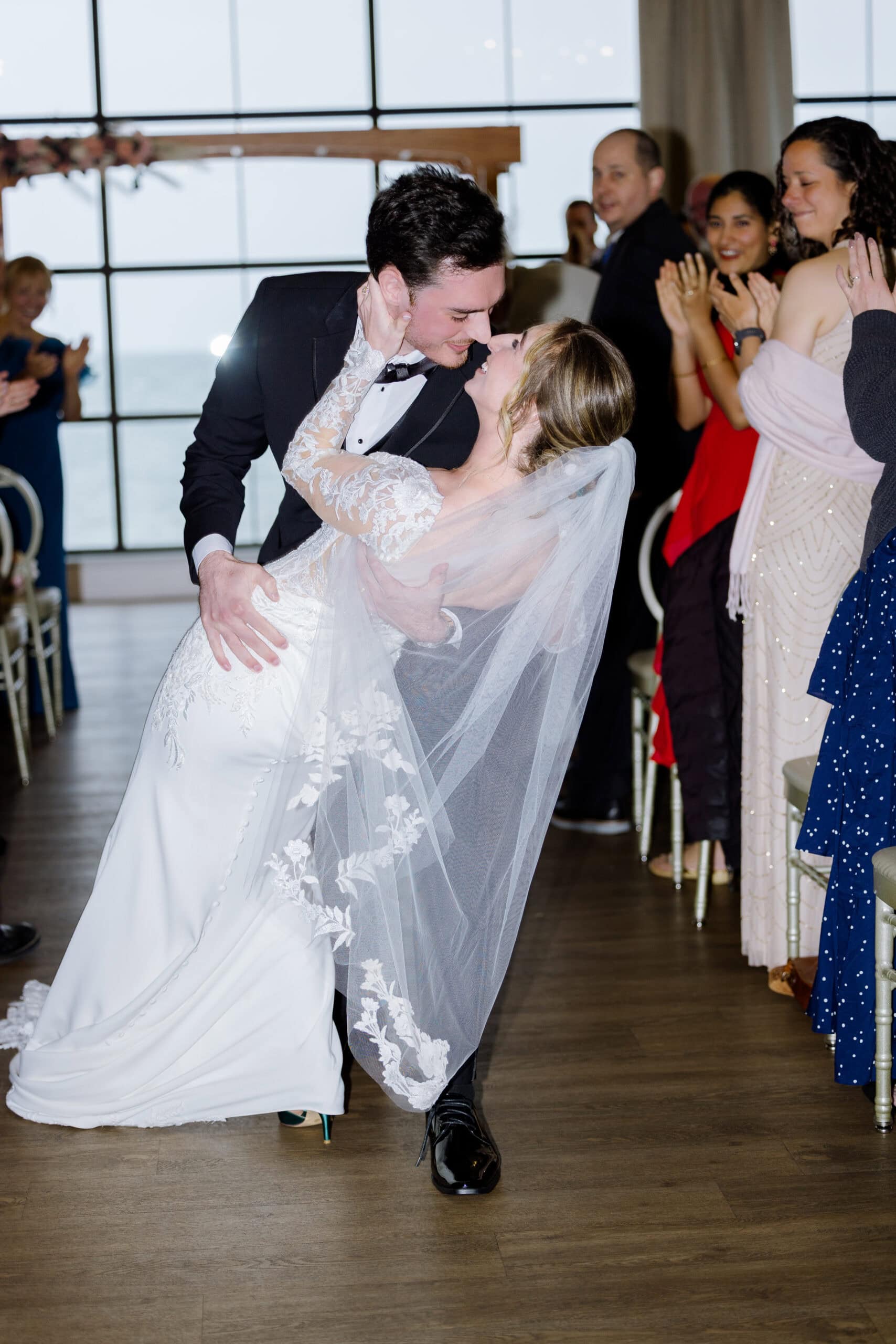 Bride in lace wedding dress and veil dips backward as groom in black tuxedo leans in for a kiss, guests clapping in background at Tyde at Walnut Beach, Milford, CT.