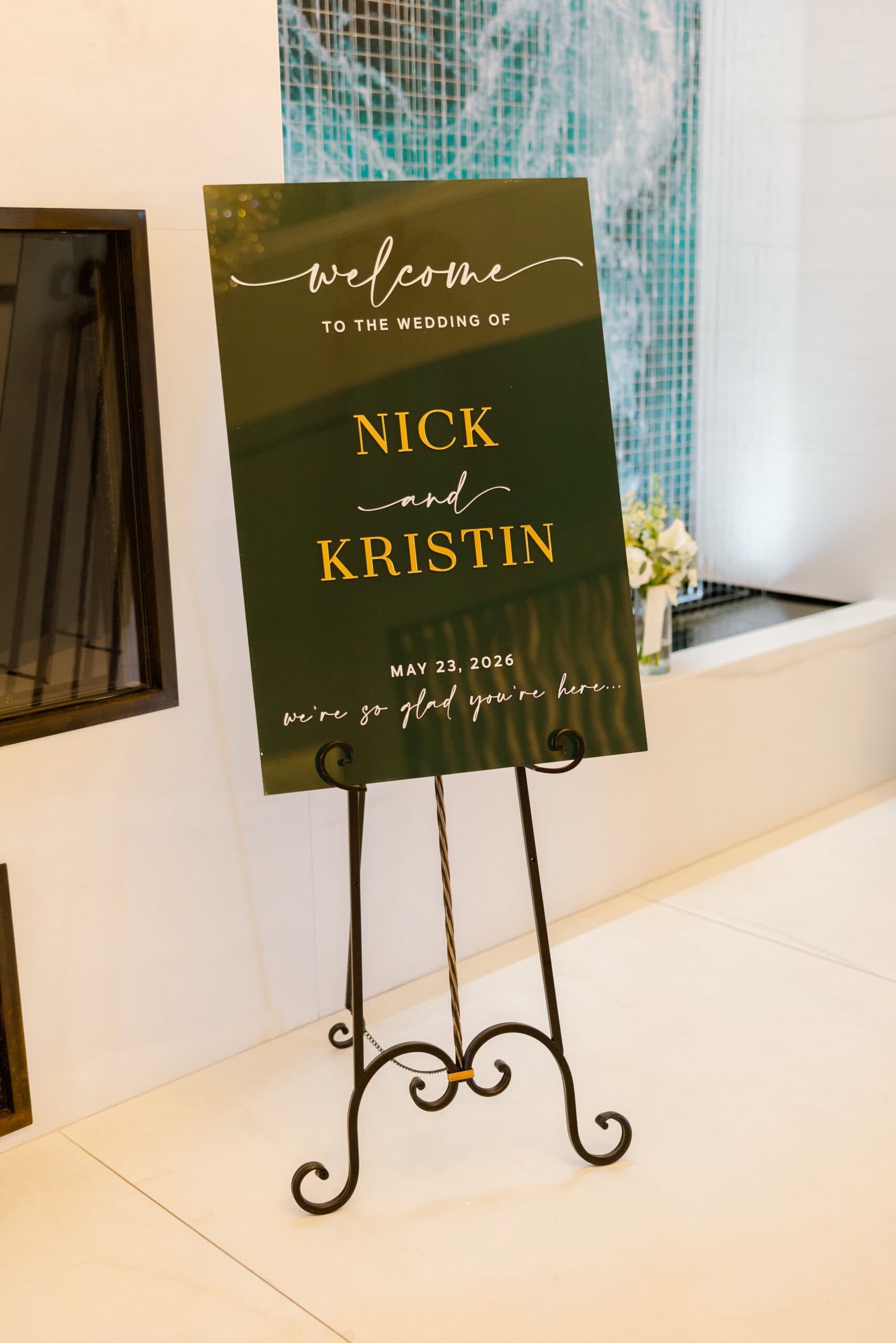 Dark green wedding welcome sign with gold and white script reading 'Welcome to the wedding of Nick and Kristin, May 23, 2026' on black wrought iron stand.