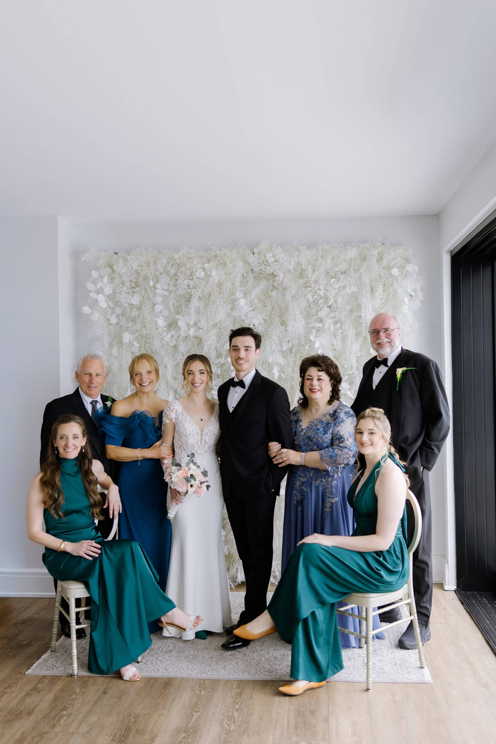 Bride and groom with family in formal attire posing indoors before a textured white floral backdrop at Tyde at Walnut Beach, Milford, CT.