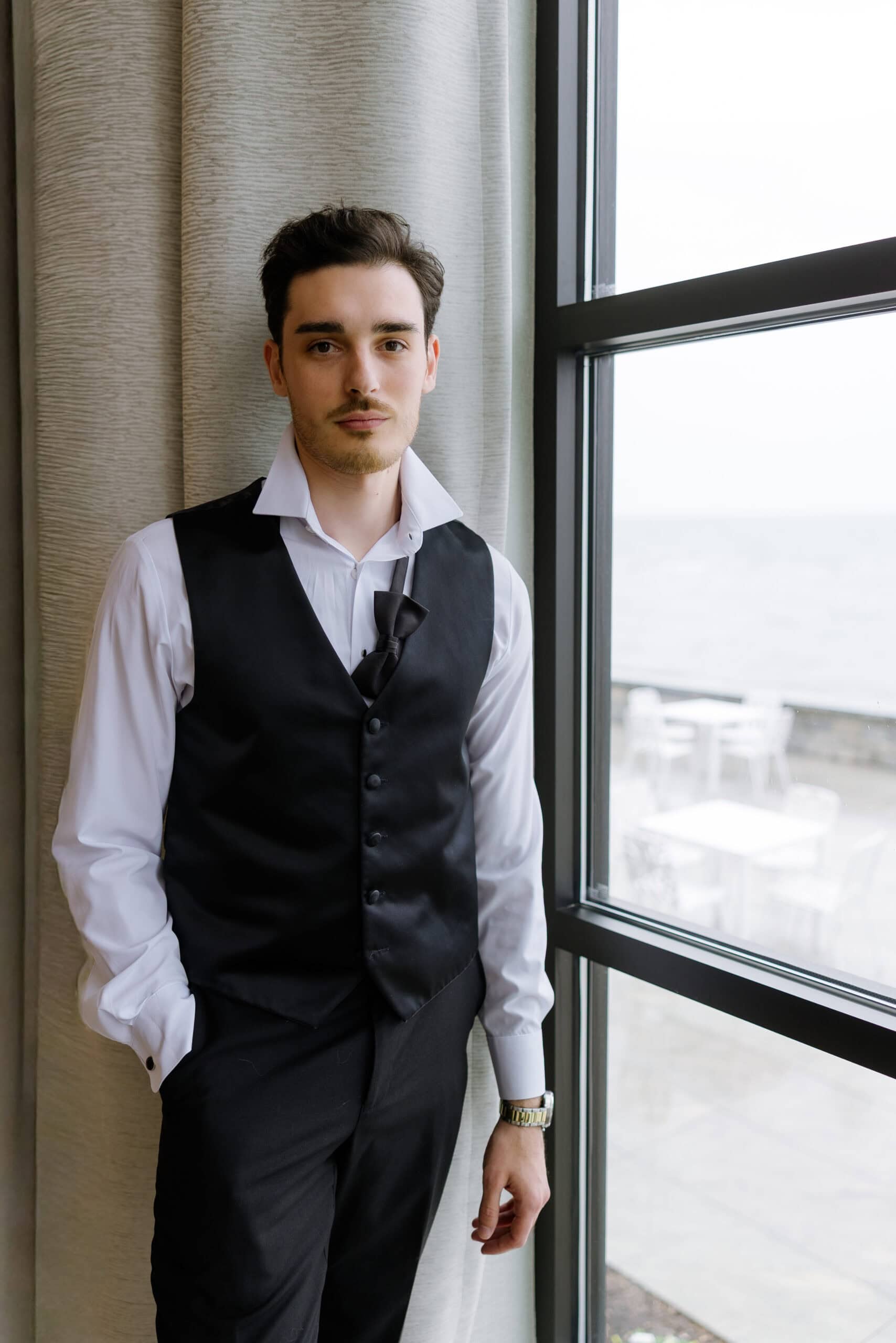 Man in white dress shirt and black waistcoat with loosened bow tie standing by window with ocean view and neutral curtains
