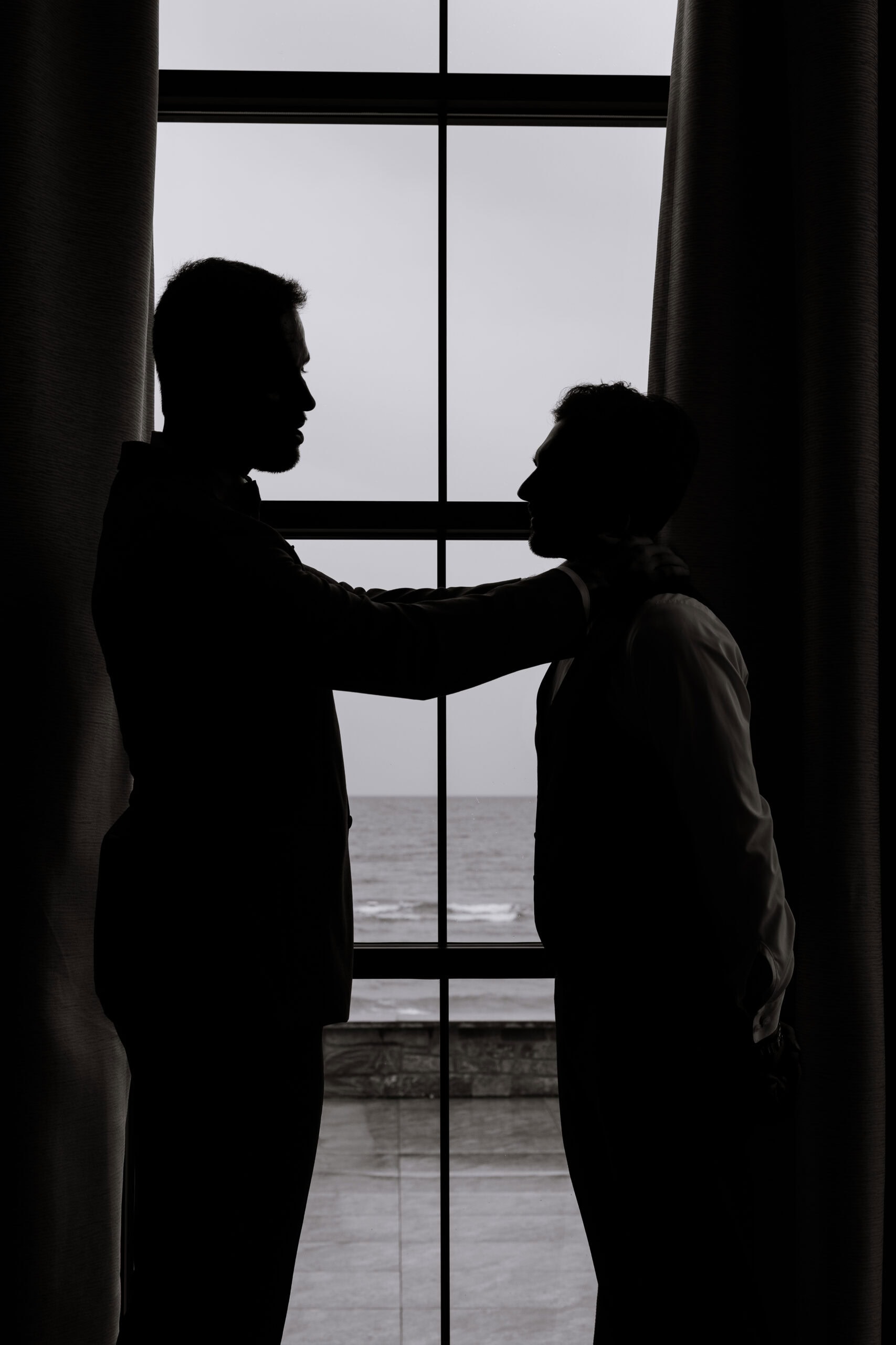Silhouette of two men facing each other by an oceanfront window with curtains, one adjusting the other's collar.