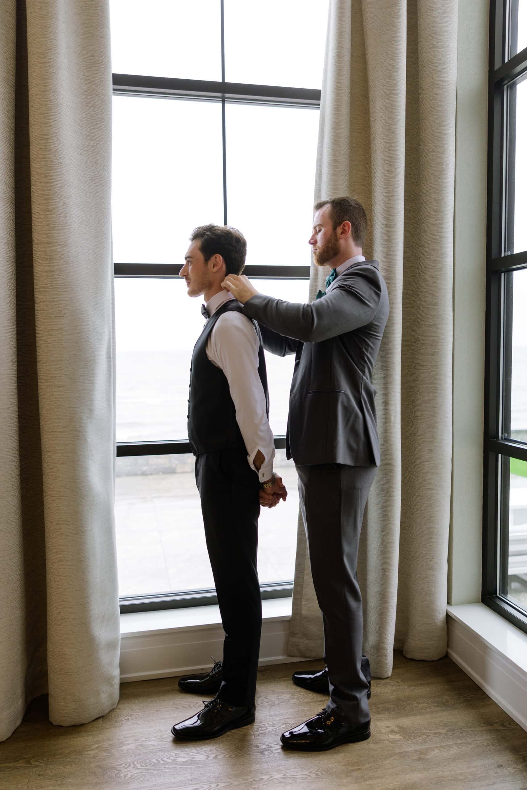Two men in formal attire by a large window with beige curtains; one adjusts the other's collar in preparation for a wedding at Tyde at Walnut Beach, Milford, CT.