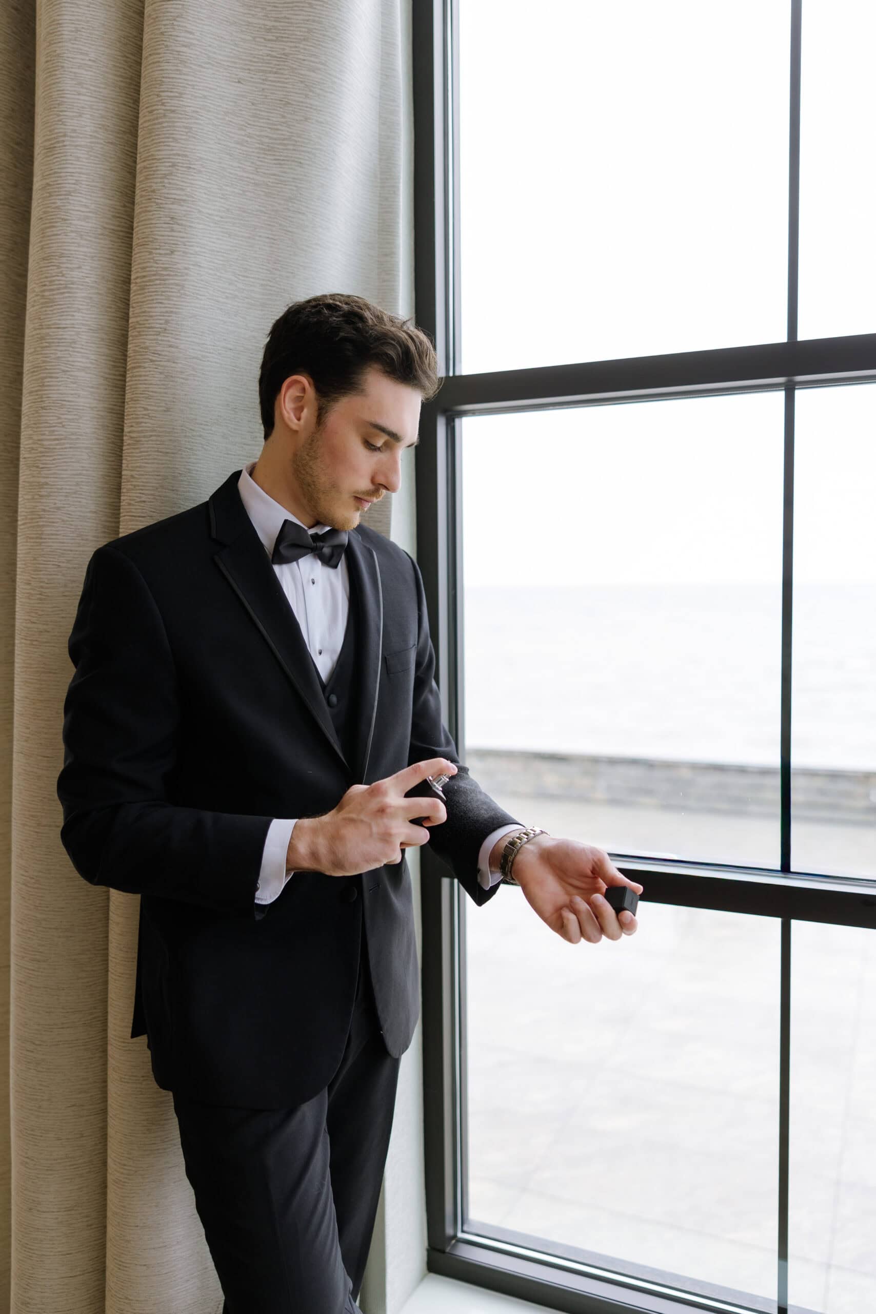 Man in black tuxedo applying cologne near window with beige curtains and soft natural light