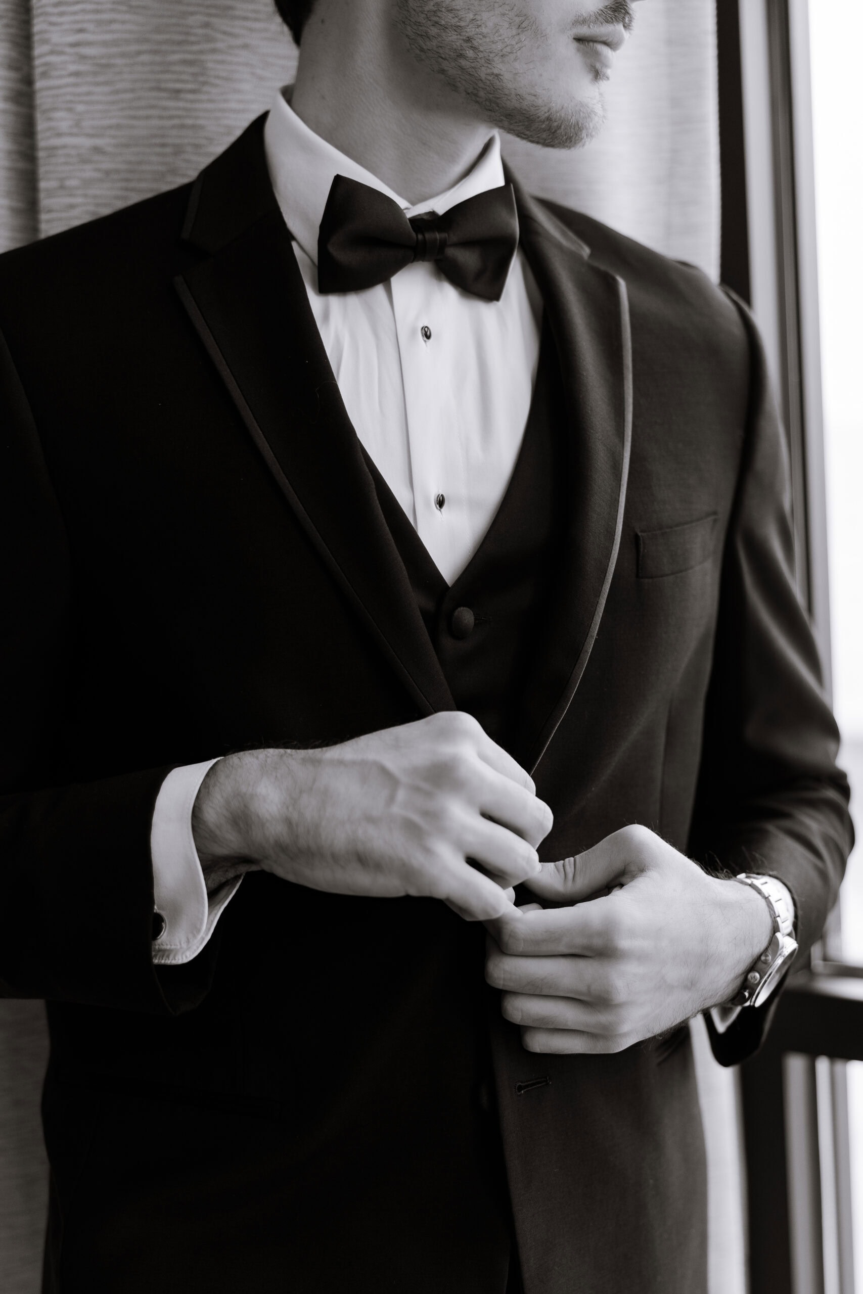 Close-up of groom buttoning black tuxedo jacket with bow tie and white shirt, standing near window at Tyde at Walnut Beach, Milford, CT.