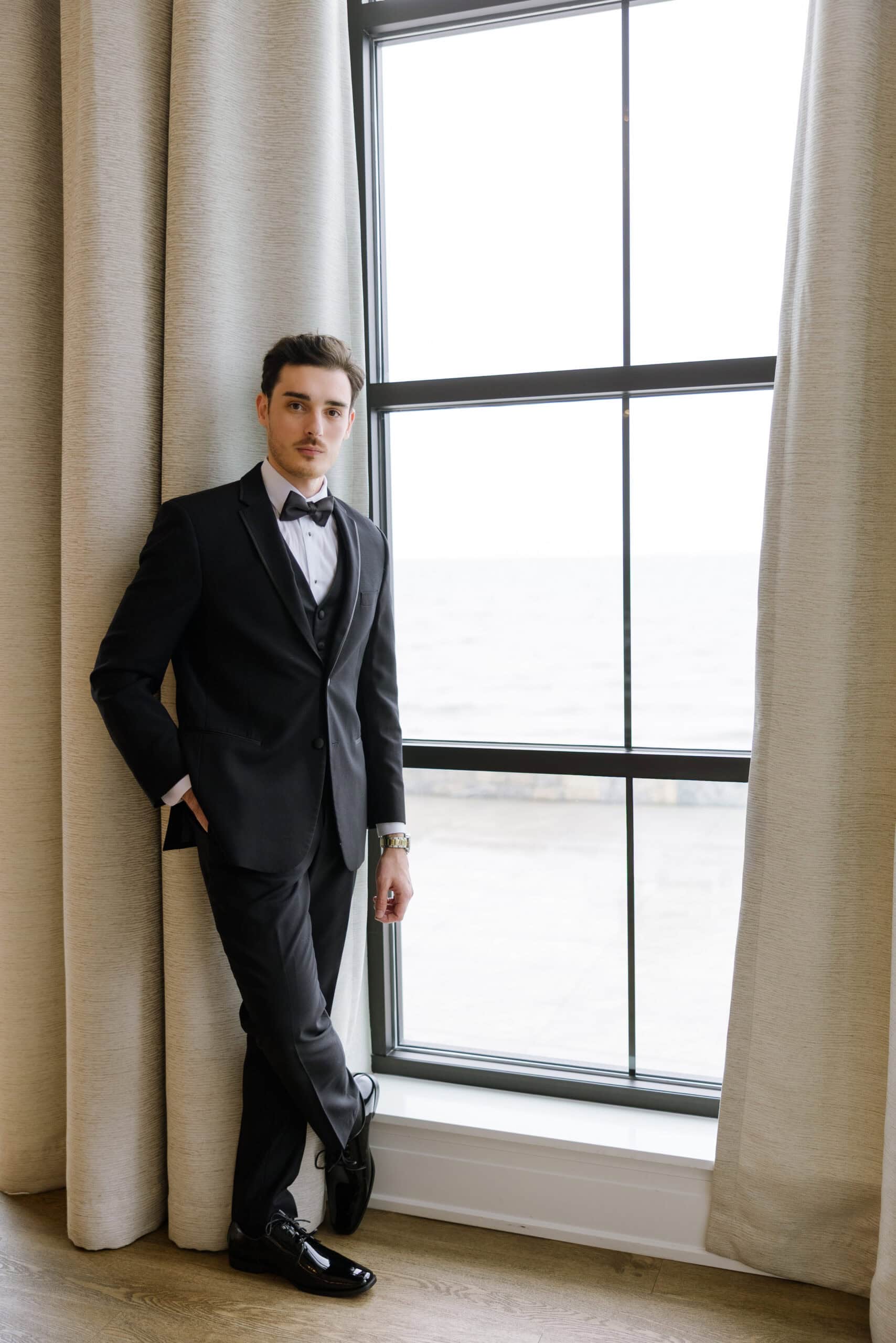 Man in black tuxedo with bow tie standing by window with beige curtains, waterfront view outside, Tyde at Walnut Beach venue