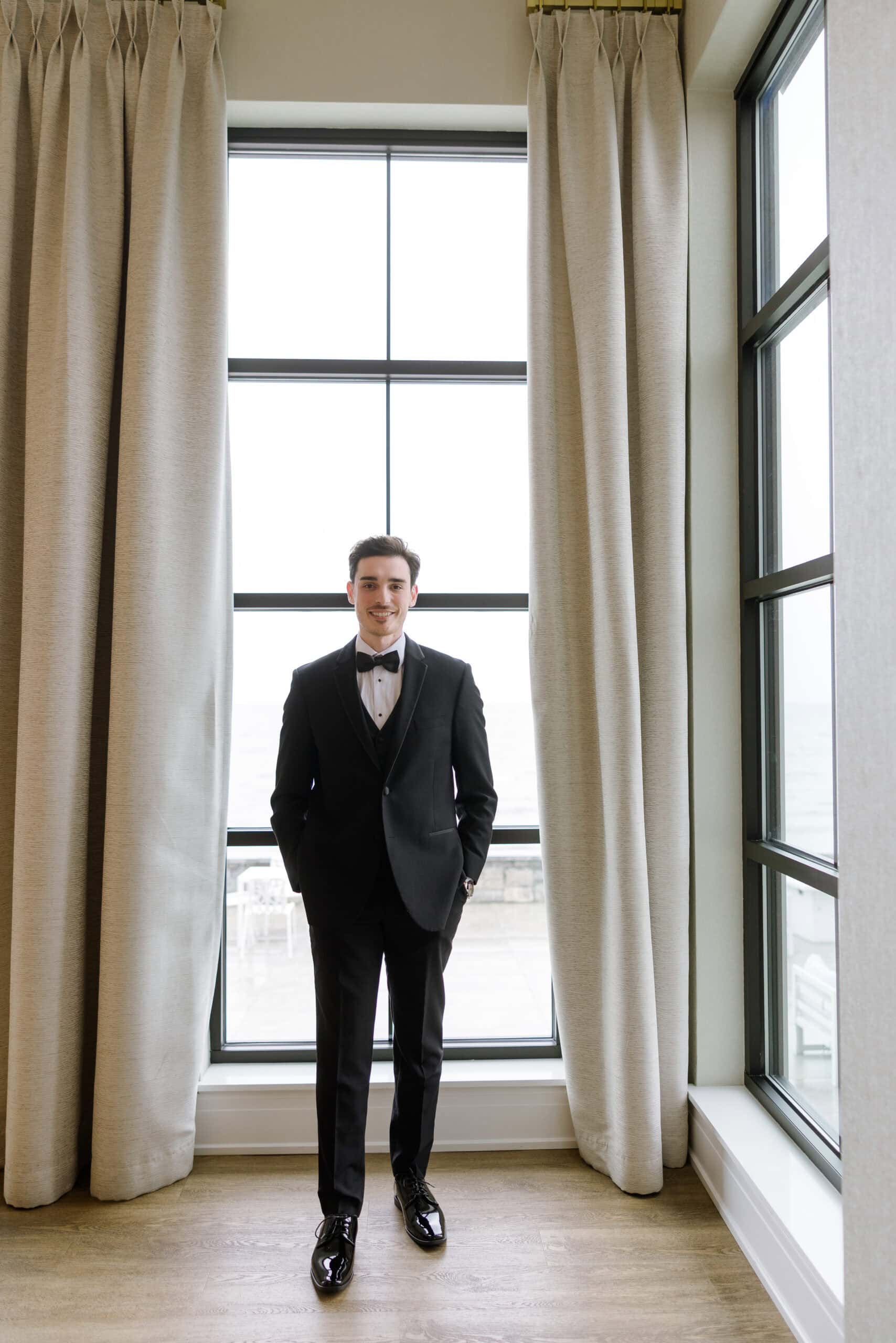 Groom in black tuxedo standing between beige curtains in front of large window with waterfront view at Tyde at Walnut Beach, Milford, CT.