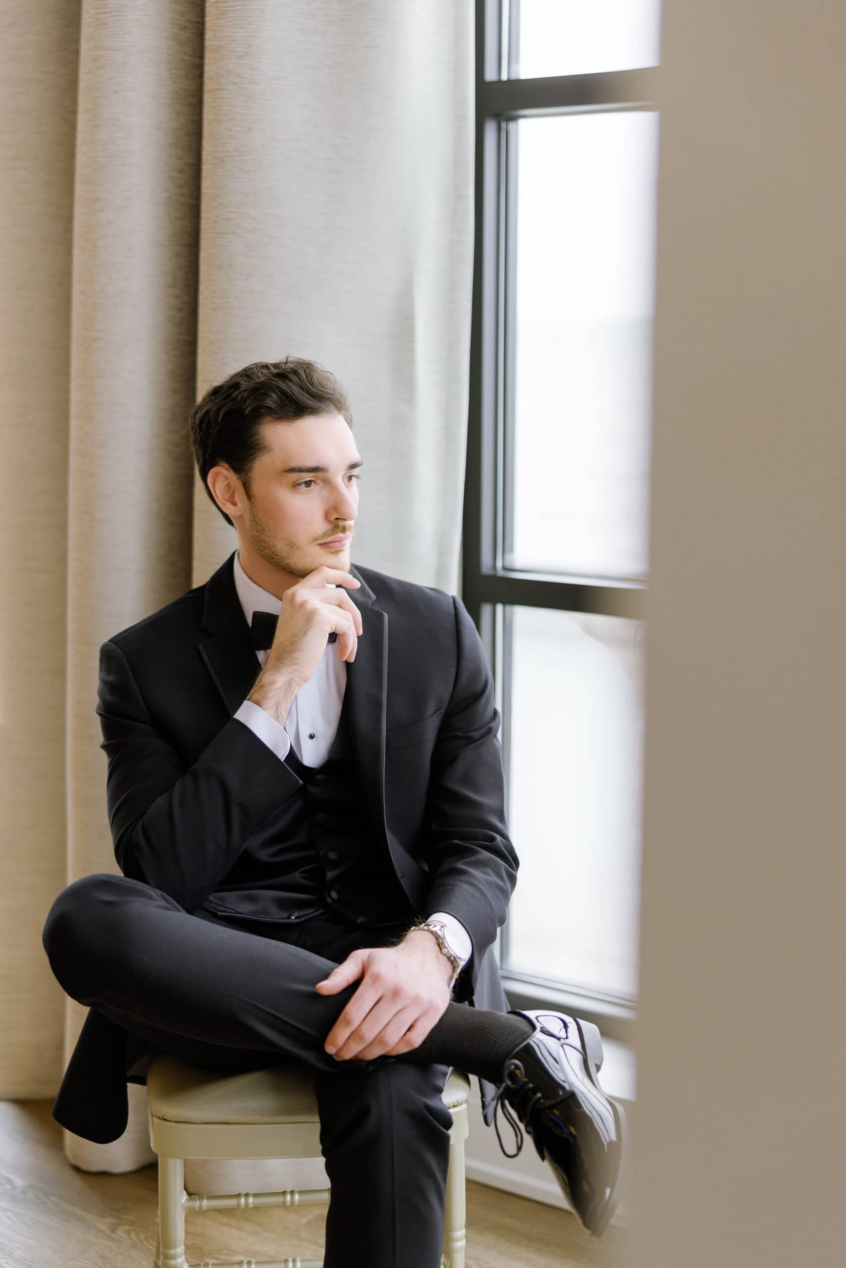 Man in black tuxedo sitting on chair by window with beige curtains, looking contemplative, hand near chin.