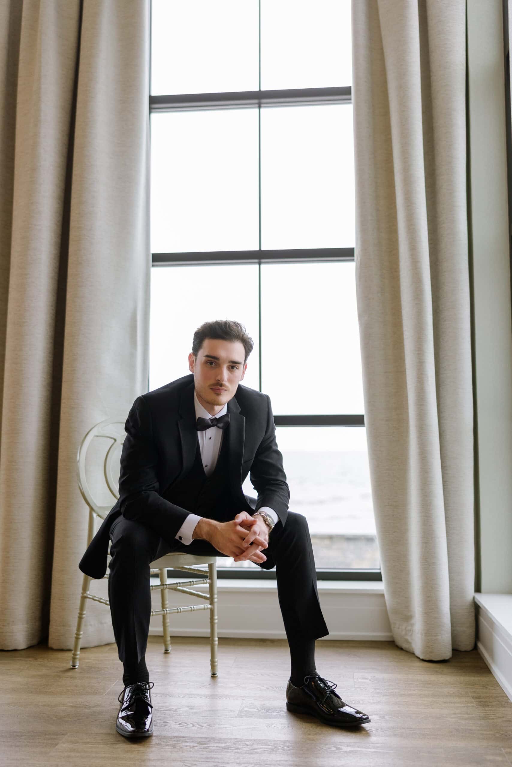 Man in black tuxedo sitting on a translucent chair in front of a large window with beige curtains at Tyde at Walnut Beach, Milford, CT.