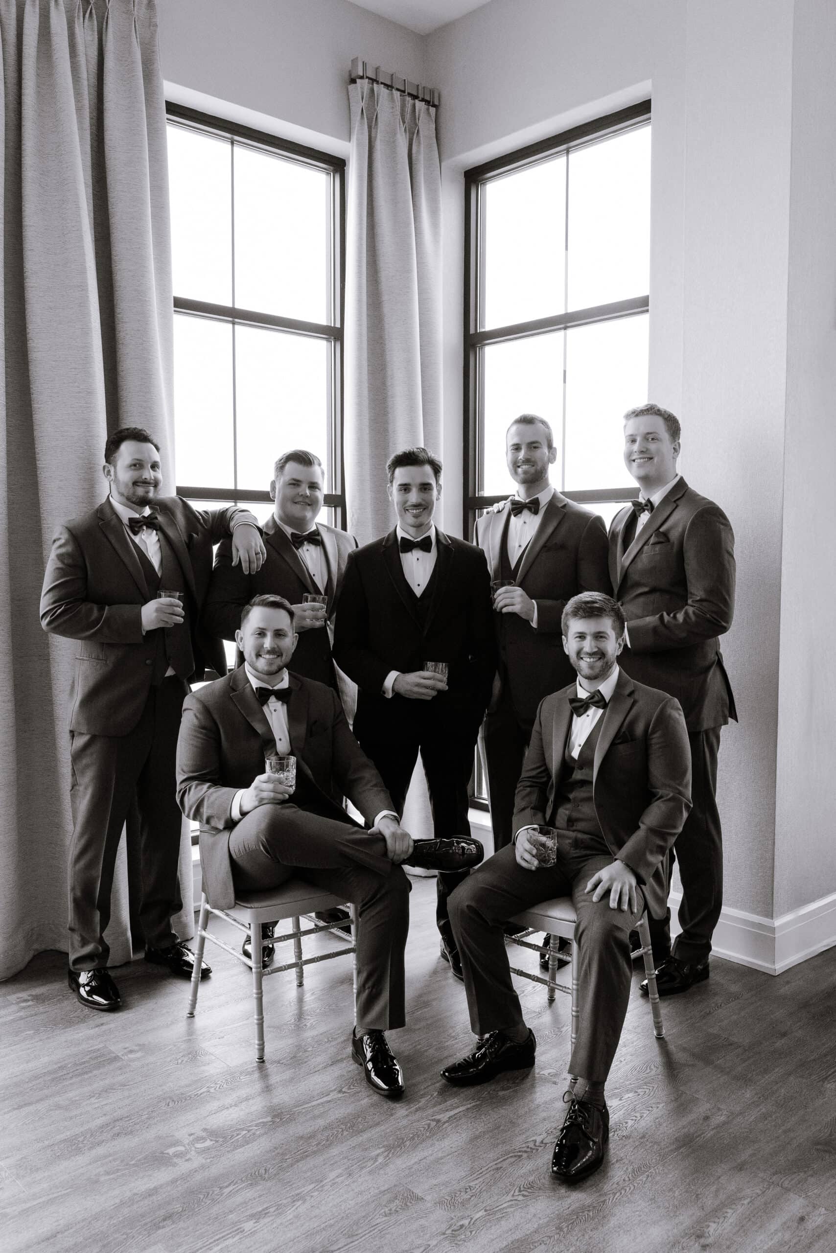Seven groomsmen in tuxedos holding drinks, posed in a corner with large windows and curtains, natural light illuminating the scene at Tyde at Walnut Beach, Milford, CT.