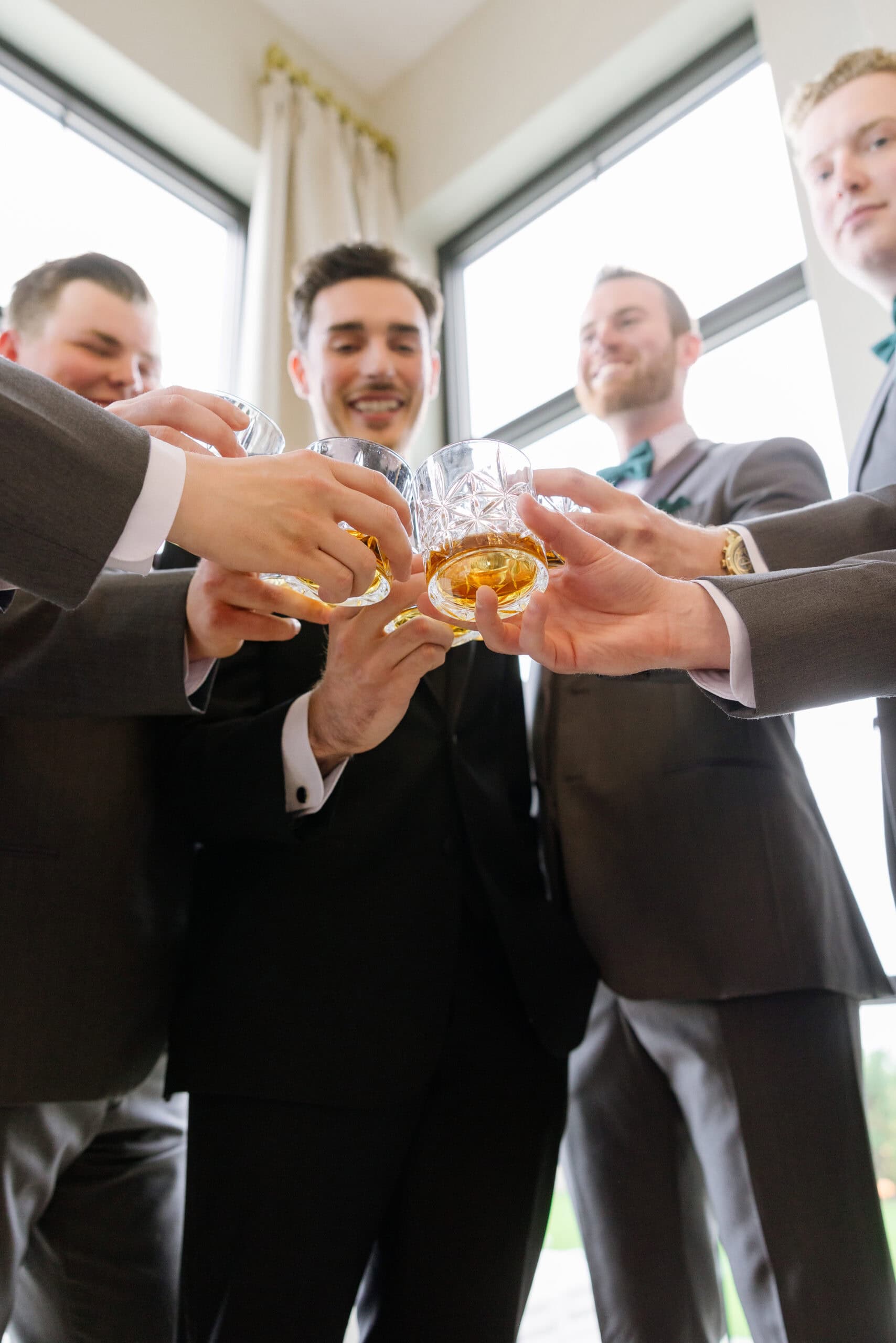 Close-up of groomsmen in suits clinking crystal glasses with amber liquid, standing near large windows with soft daylight.