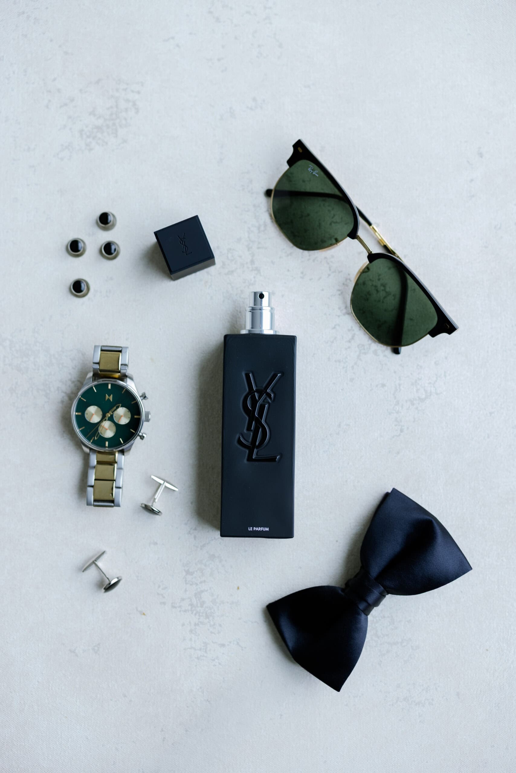 Flat lay of groom's accessories including YSL Le Parfum bottle, Ray-Ban sunglasses, two-tone chronograph watch, black onyx cufflinks, and black silk bow tie on light textured surface.