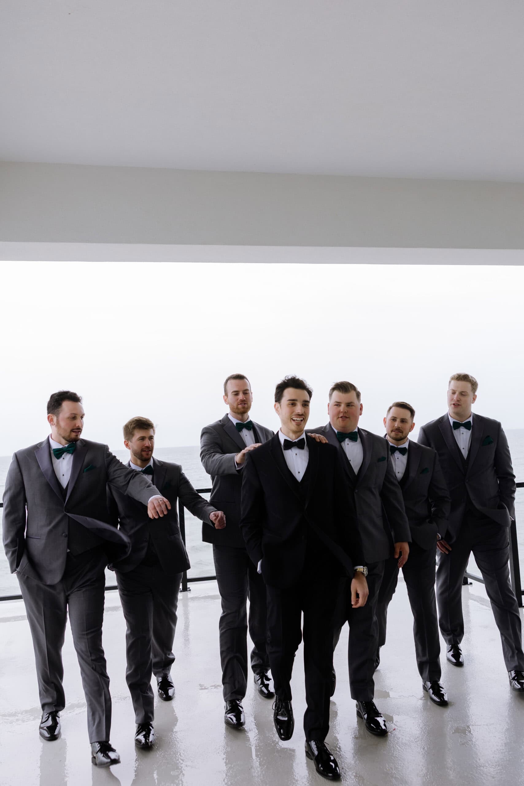 Seven men in tuxedos with green bow ties and pocket squares walking together on a covered terrace overlooking water at Tyde at Walnut Beach, Milford, CT.
